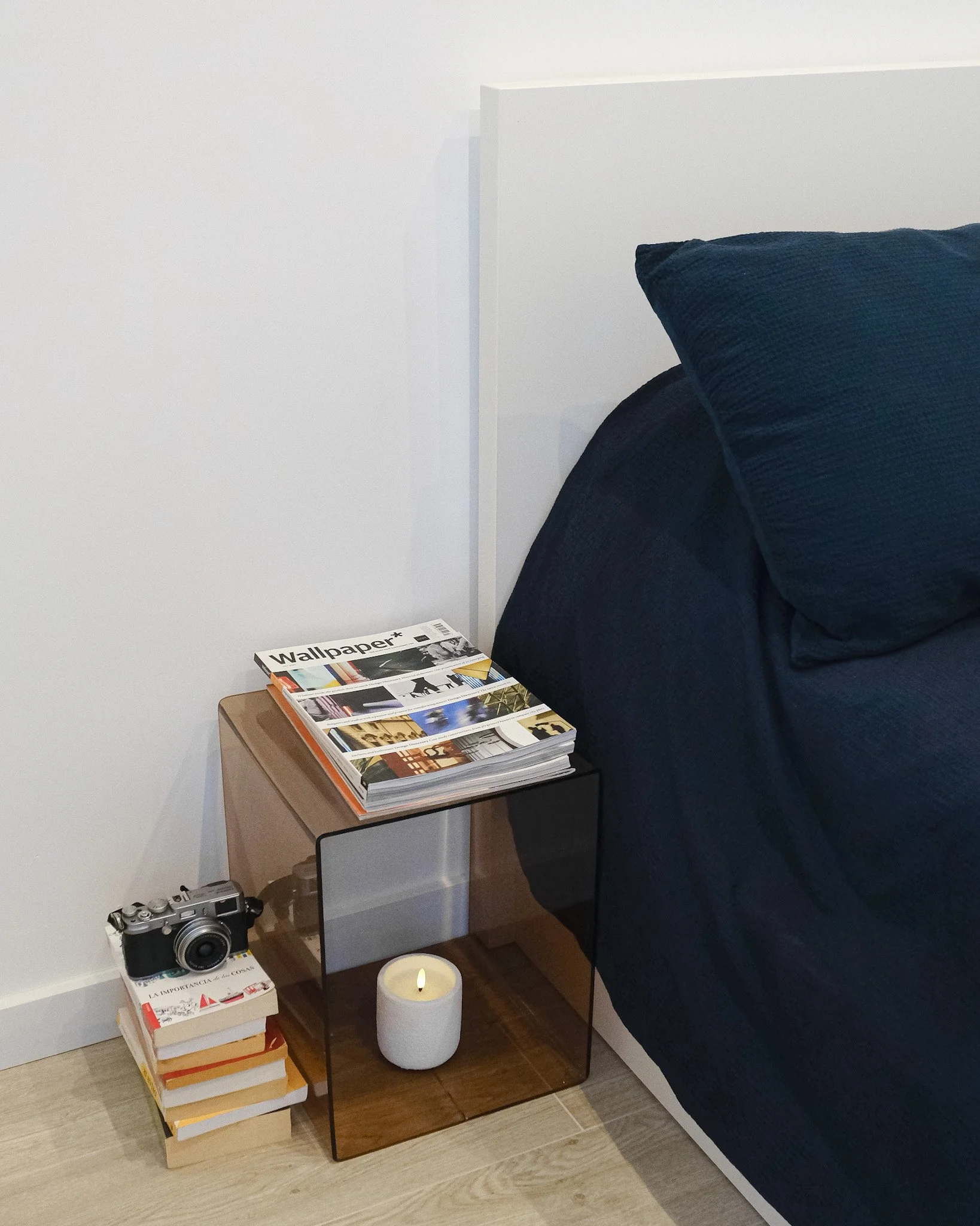 Mesita de noche con revistas, cámara antigua, libros y una vela encendida, junto a una cama con sábanas oscuras.