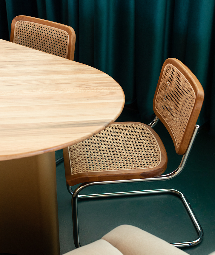 Sillas de madera con respaldo de cestería junto a mesa de madera redonda, cortinas verdes de fondo.