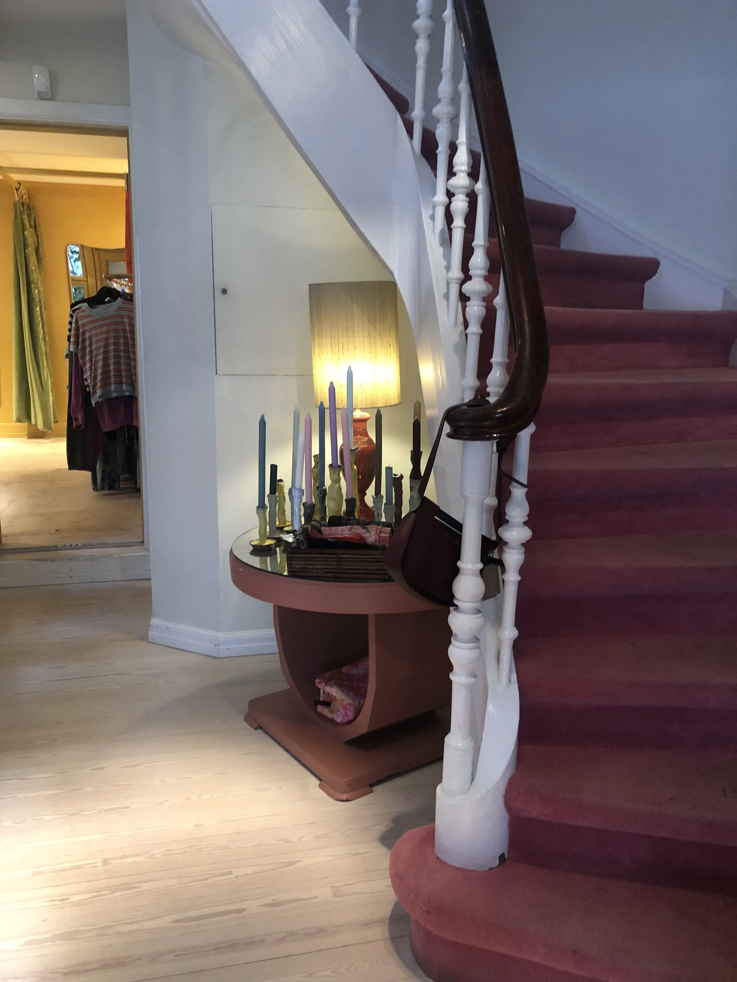 Interior de una casa con una escalera de caracol, una mesa con candelabros y una lámpara. En el fondo se ven ropa y cortinas.