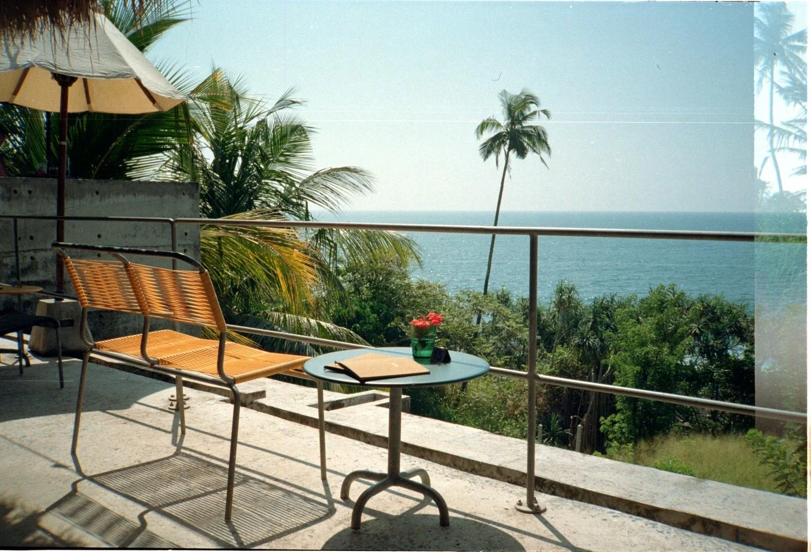 Decoración de terraza con vista al océano, incluyendo mesa con libro y flores, sillas de mimbre, sombrilla y palmeras.