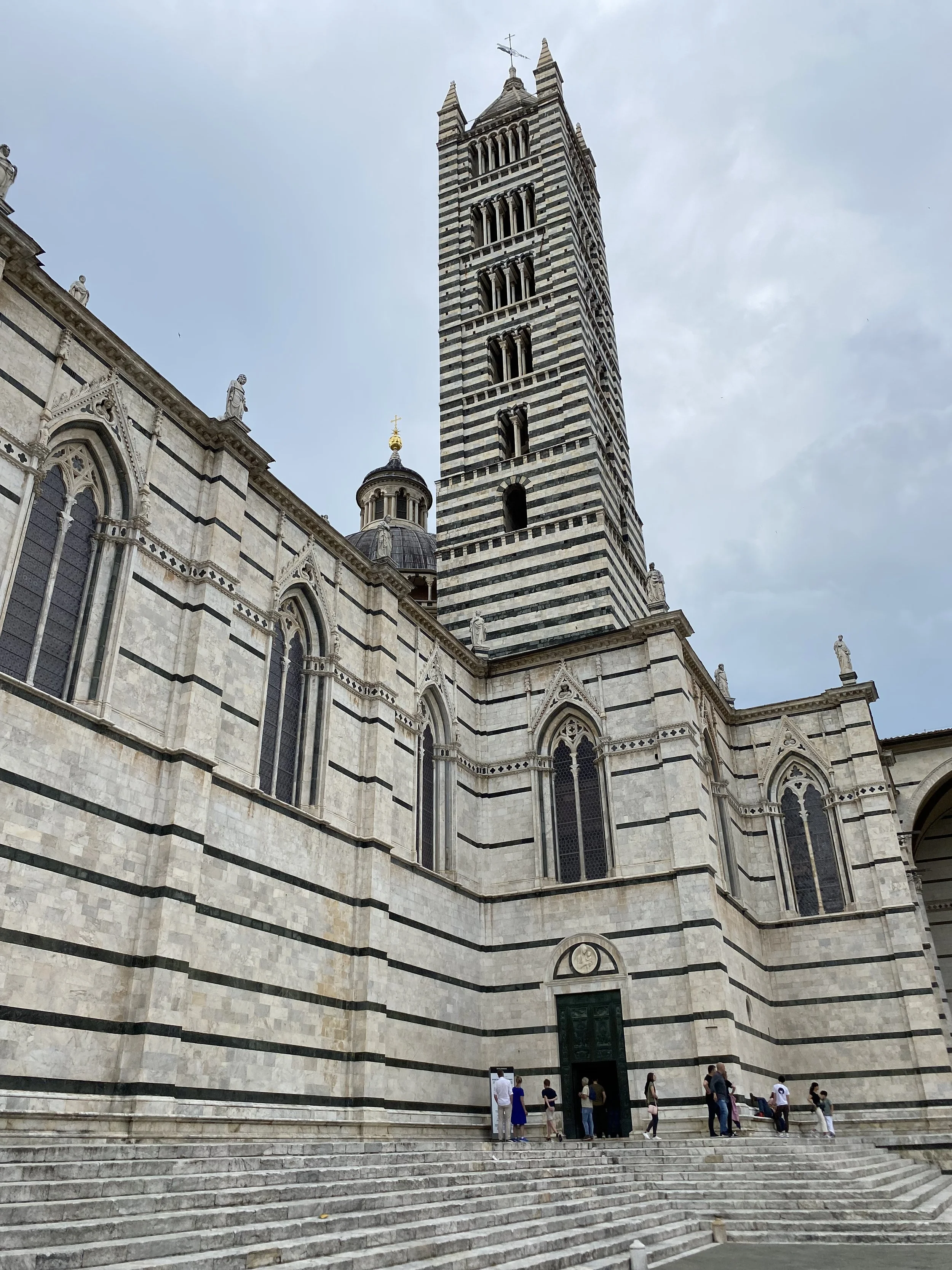 Vista de la fachada de una catedral con una torre alta con bandas blancas y negras, y varias ventanas góticas. Algunas personas están en la entrada.