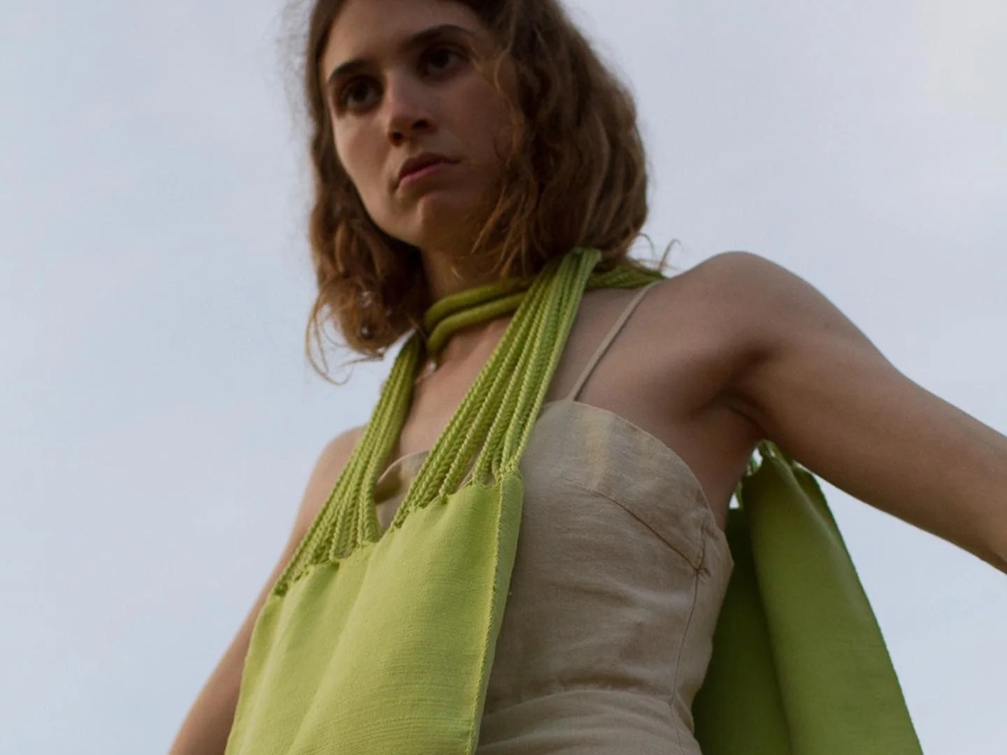 Mujer con cabello rizado, vestida con un vestido beige y rodeada de telas verdes en un fondo de cielo despejado.