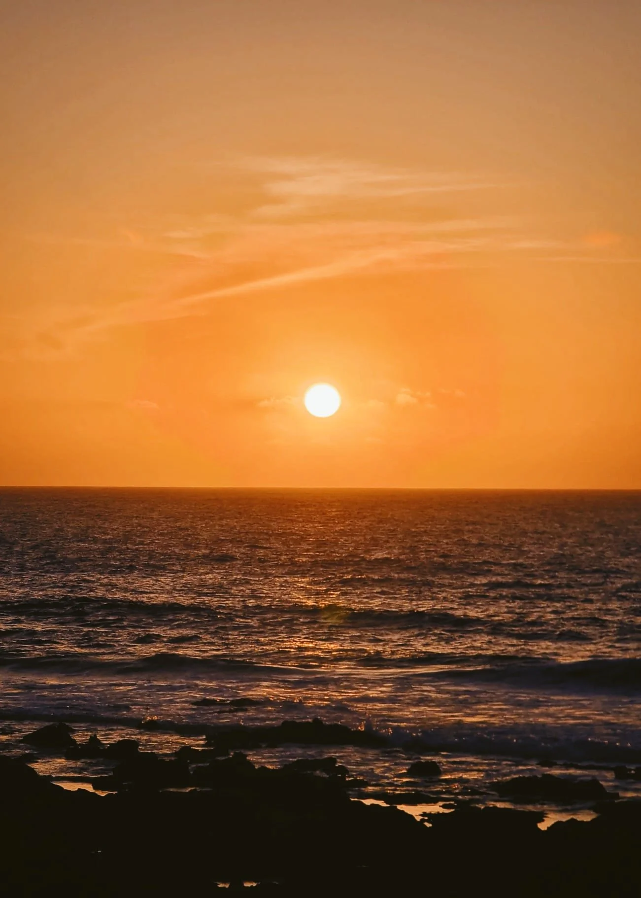 Atardecer en la playa con el sol brillante y el cielo anaranjado, olas en el mar y rocas en primer plano.