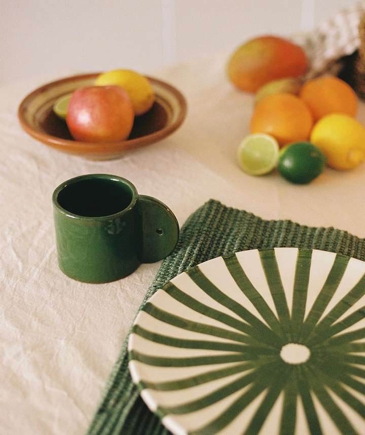 Taza verde, plato con rayas verdes y blancas, frutas (manzana, limón, durazno, lima, naranja), manteles y fondo decorativo.