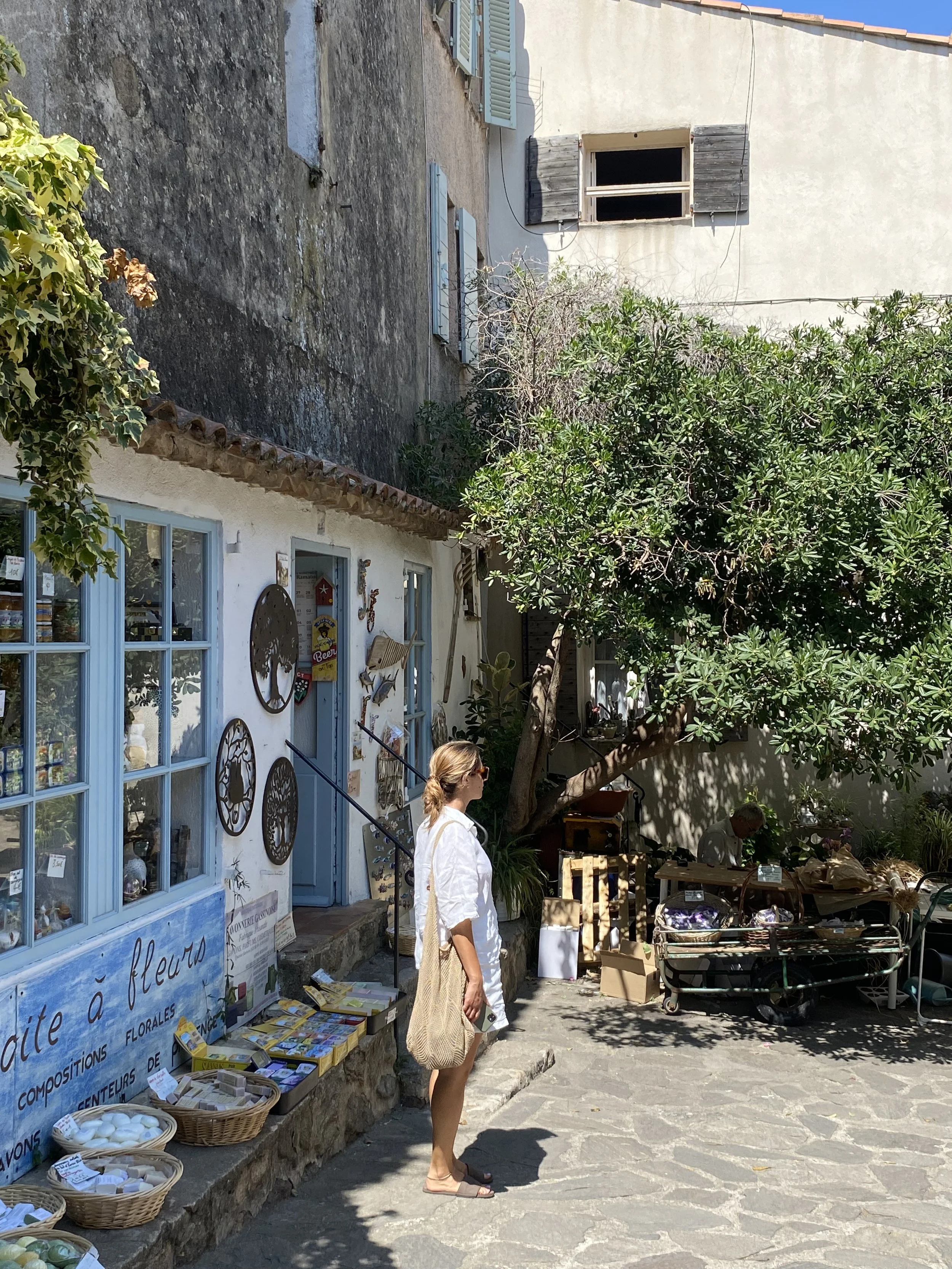 Mujer de perfil con camiseta blanca, pantalones cortos y mochila de yute, de pie en una calle adoquinada con tienda de flores y decoración rústica, rodeada de árboles y otras personas en un día soleado.