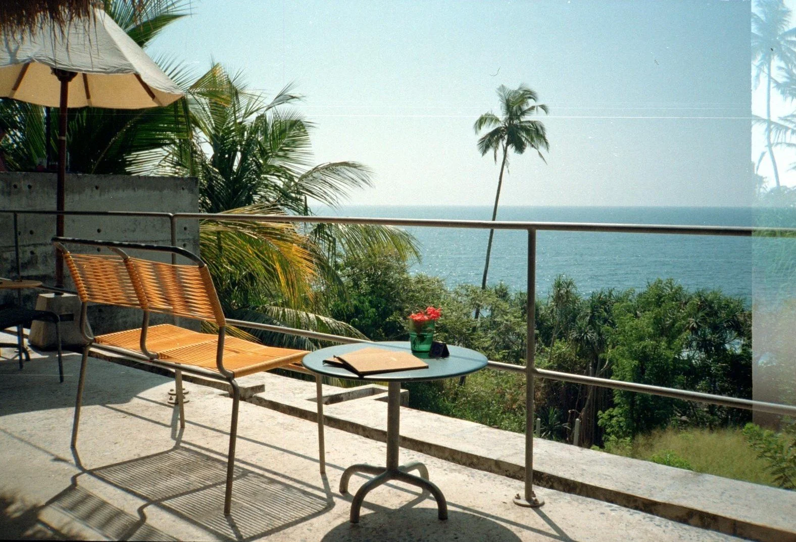 Vista de una terraza con sillas de mimbre, una mesa pequeña con un libro, un florero con flores y vista al mar y palmeras en un día soleado.