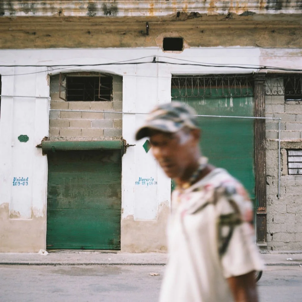 Persona con camisa y gorra en primer plano, fondo de una edificación con puertas de color verde y ventanas protegidas por rejas, en un ambiente urbano sencillo.