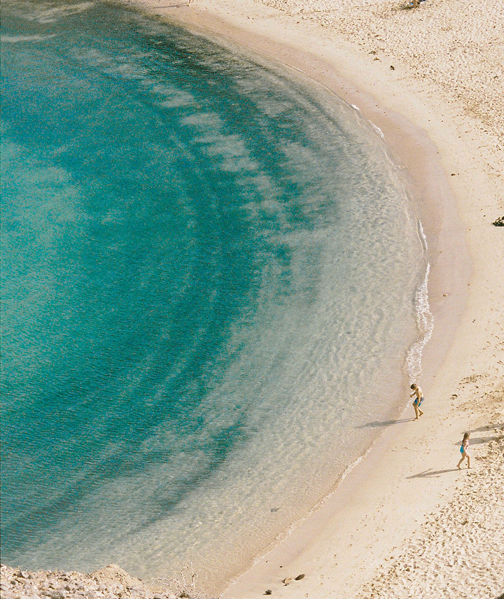 Vista aérea de una playa con agua azul y arena blanca, con dos personas caminando por la orilla.