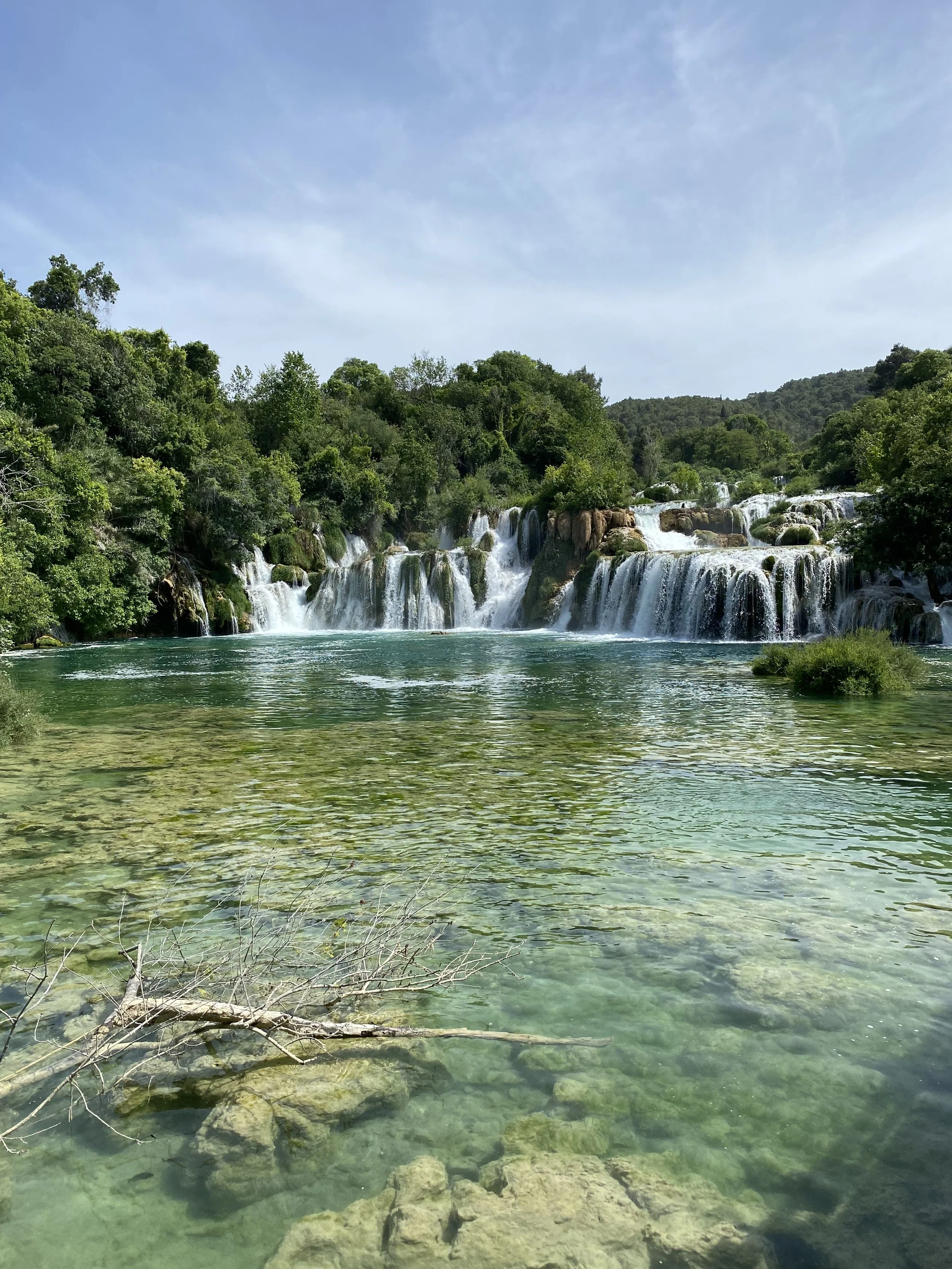 Cascada en medio de un bosque con agua clara y verde, y cielo despejado.