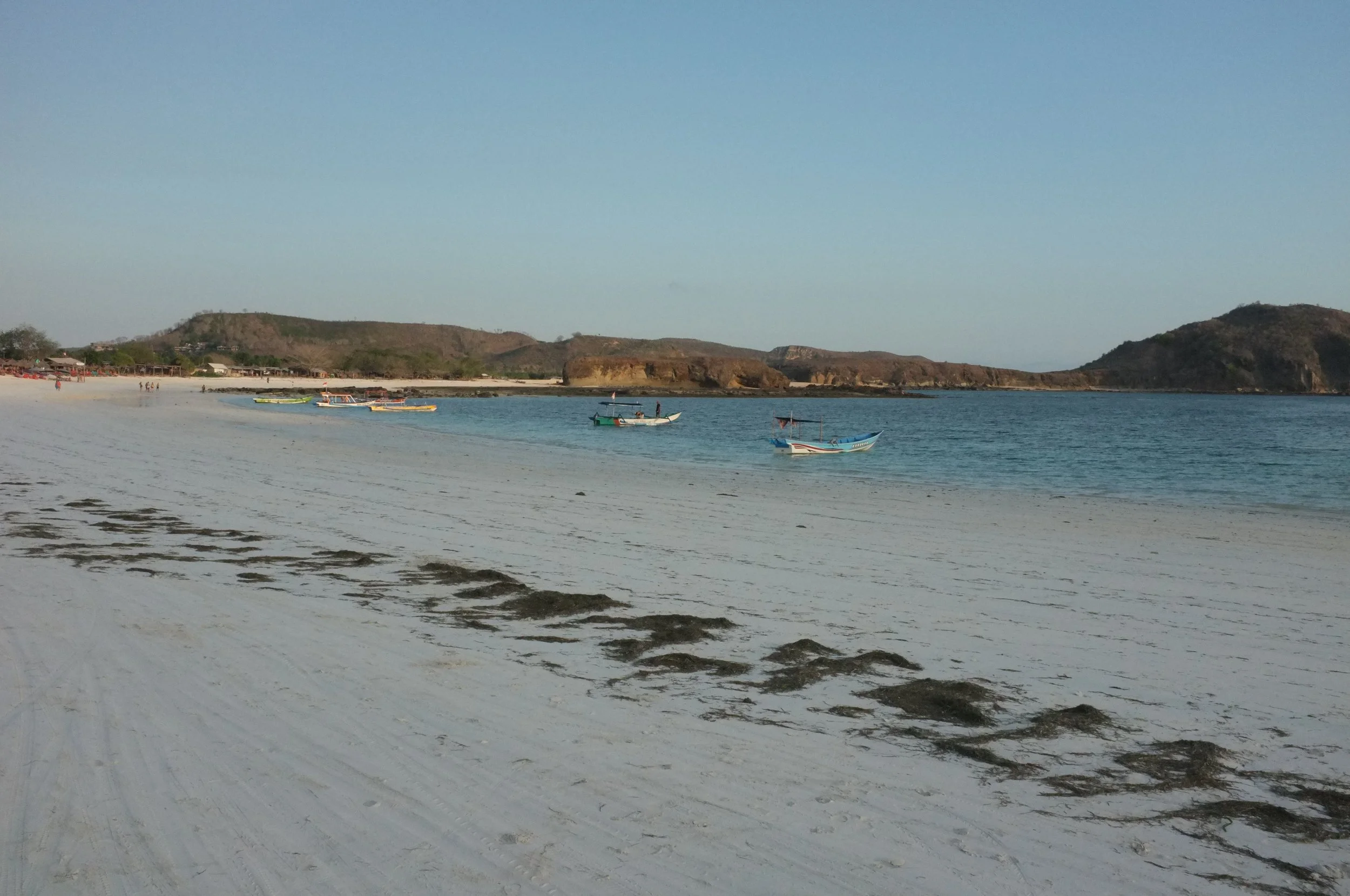 Playa con arena blanca, varias embarcaciones pequeñas en el agua, colinas y un cielo despejado.