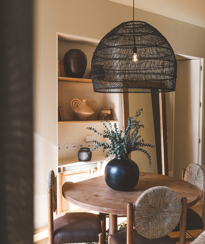Interior de una sala de comedor con mesa de madera y sillas tapizadas, un jarrón negro con plantas verdes, una lámpara colgante de ratán y una estantería con objetos decorativos.