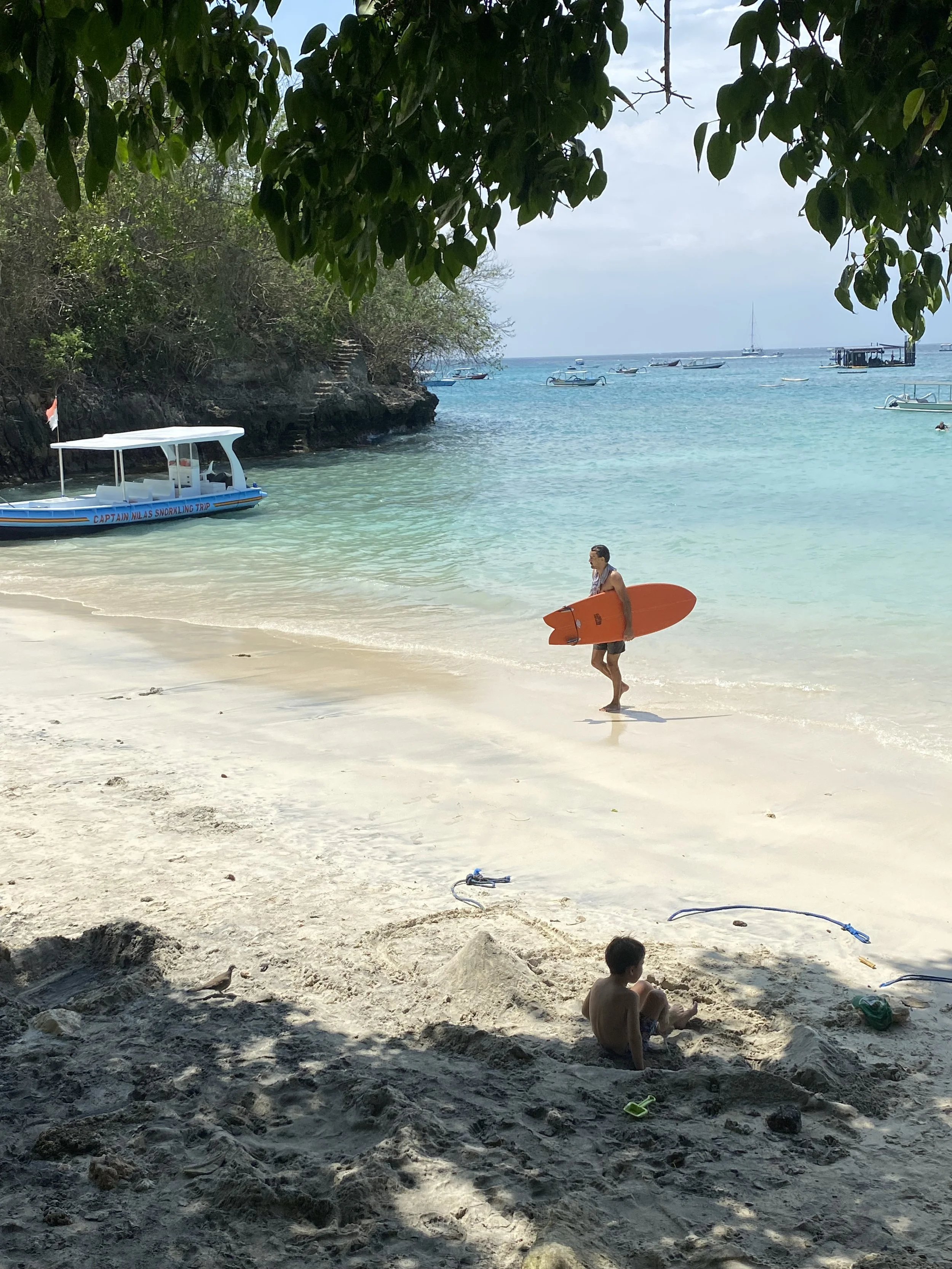 Una playa con agua cristalina, un hombre con una tabla de surf naranja entrando al mar, un niño en la arena con juguetes y barcos en el agua en el fondo.