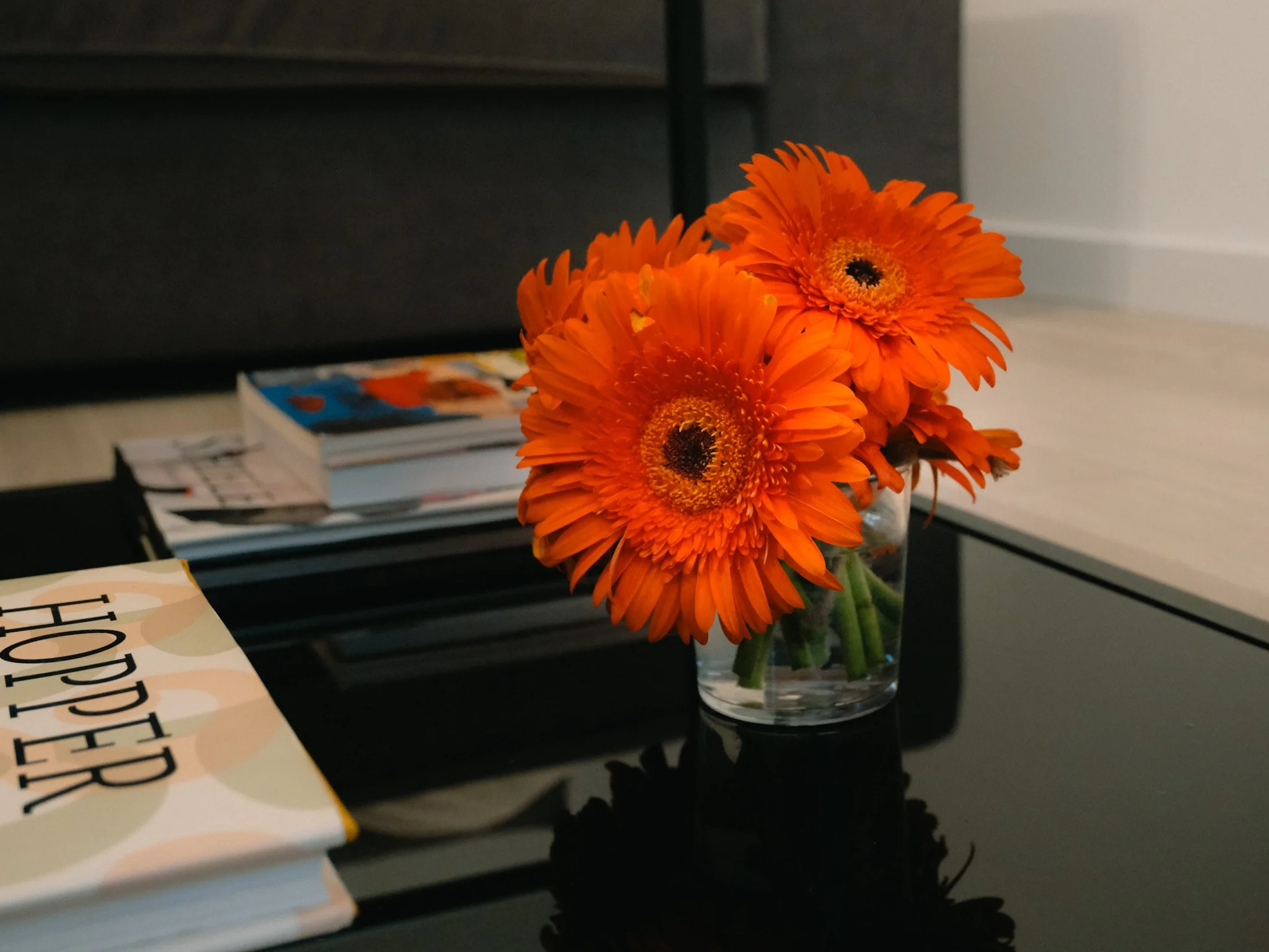 Un florero con tres flores naranjas en una mesa de escritorio, junto a unas revistas y un libro que dice 'Hopper'