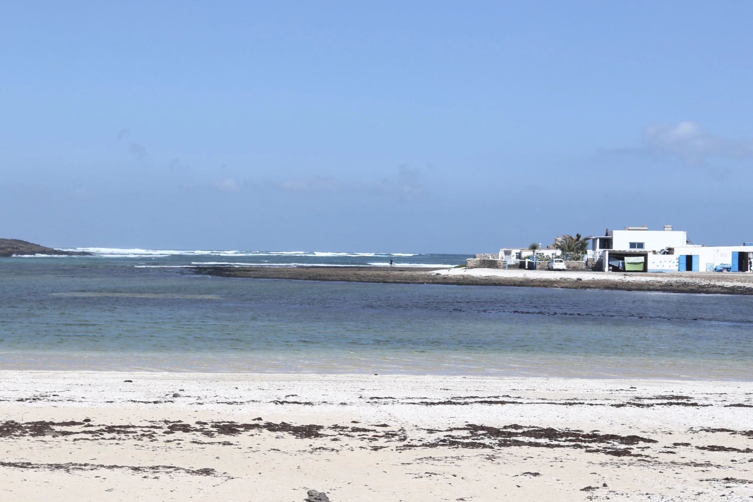 Playa con arena clara, agua tranquila y casas blancas en el fondo bajo cielo azul con algunas nubes.