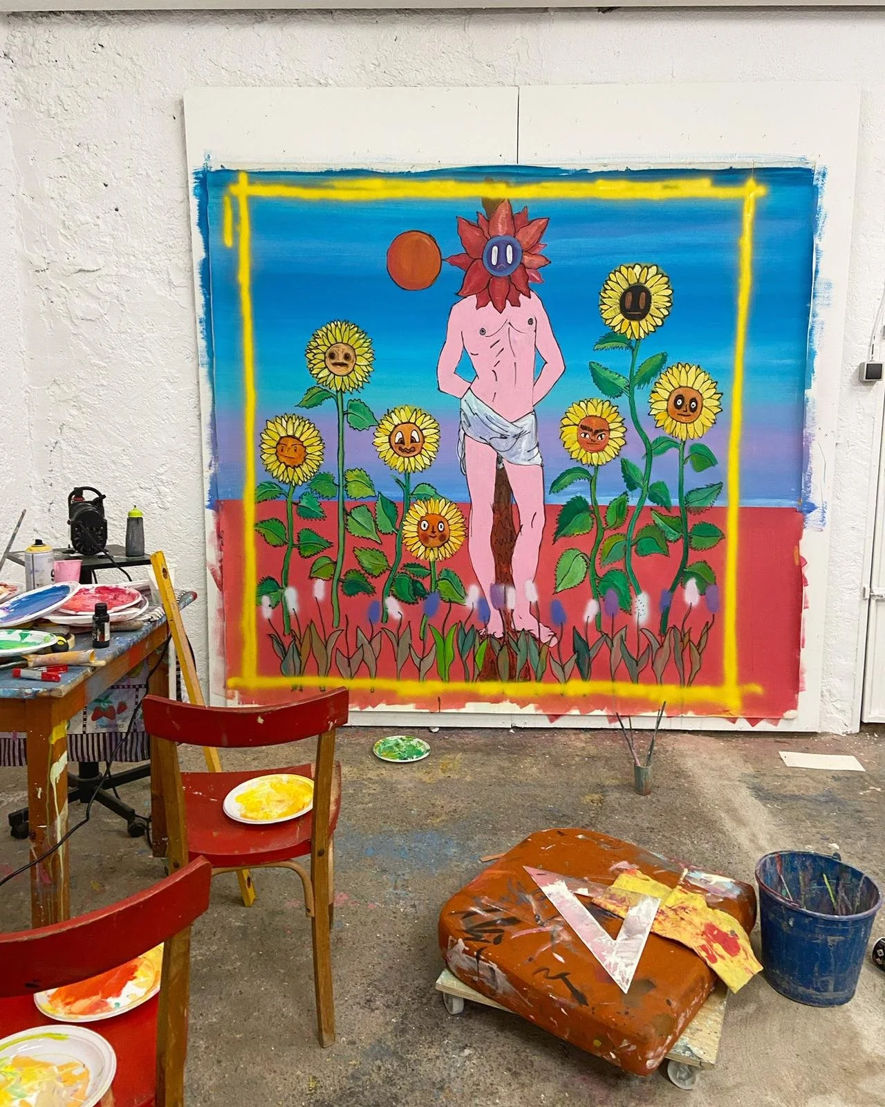 Una pintura de un personaje humano con cara de flor y cuerpo desnudo, rodeado de girasoles con caras sonrientes en un fondo de cielo azul y tierra roja, en un taller de arte.