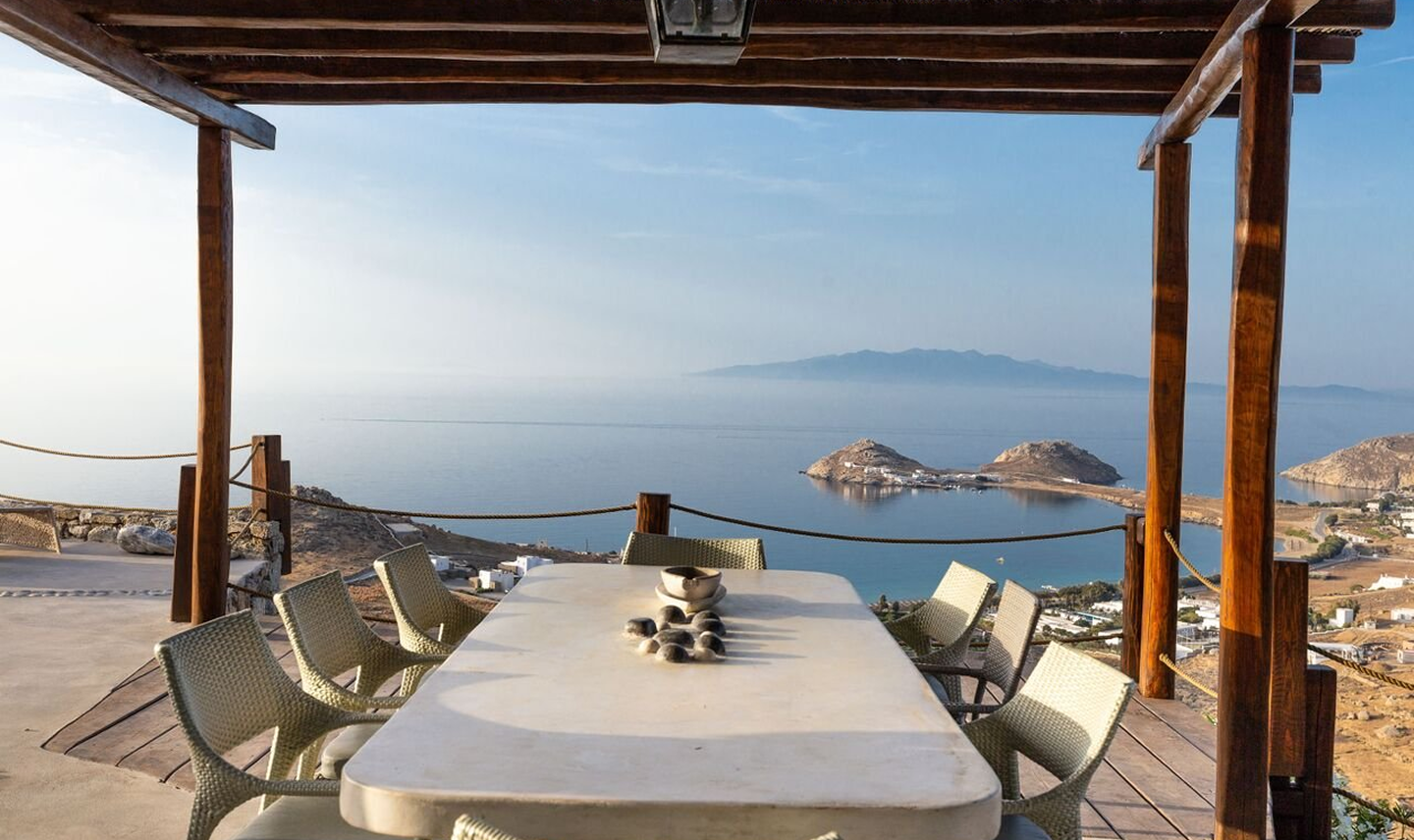 Mesa de comedor con siete sillas en un balcón con vista al mar y a islas en un paisaje costero
