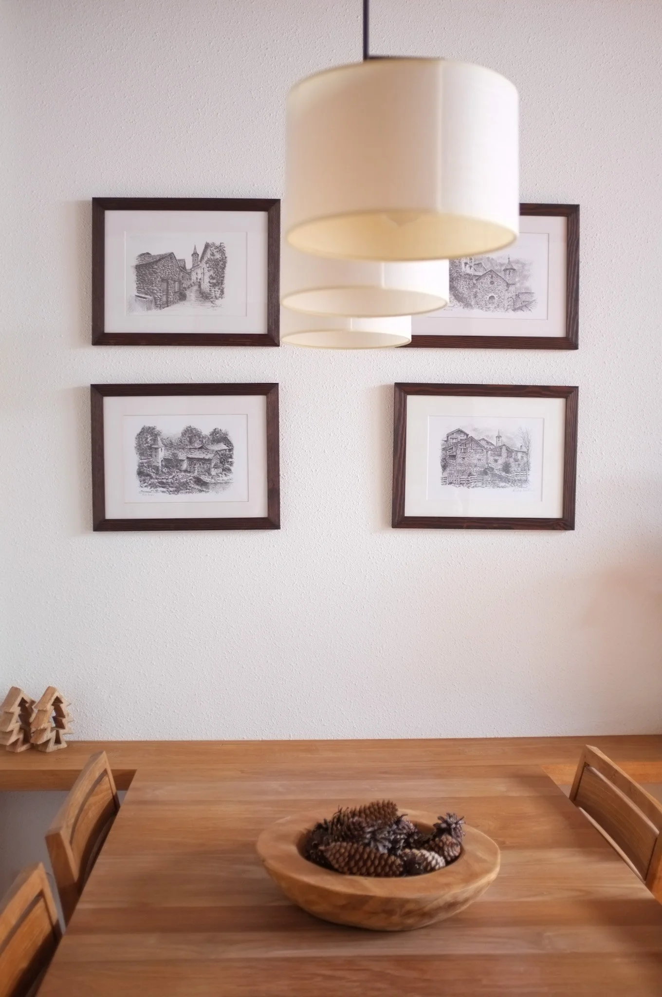 Plano de una pared con cuatro cuadros enmarcados con dibujos de escenas rurales, colgante de lámpara blanca y mesa de madera con un plato de piñas y decoración navideña.
