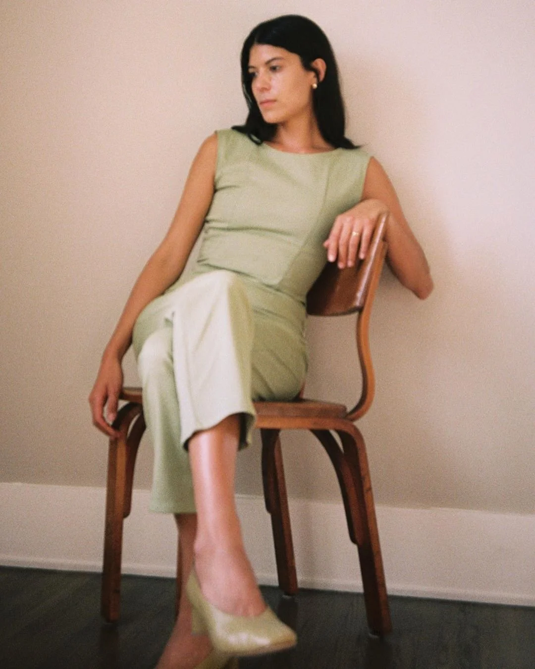 Mujer joven con vestido verde sentada en una silla de madera, mirando hacia la izquierda, con fondo de pared clara.