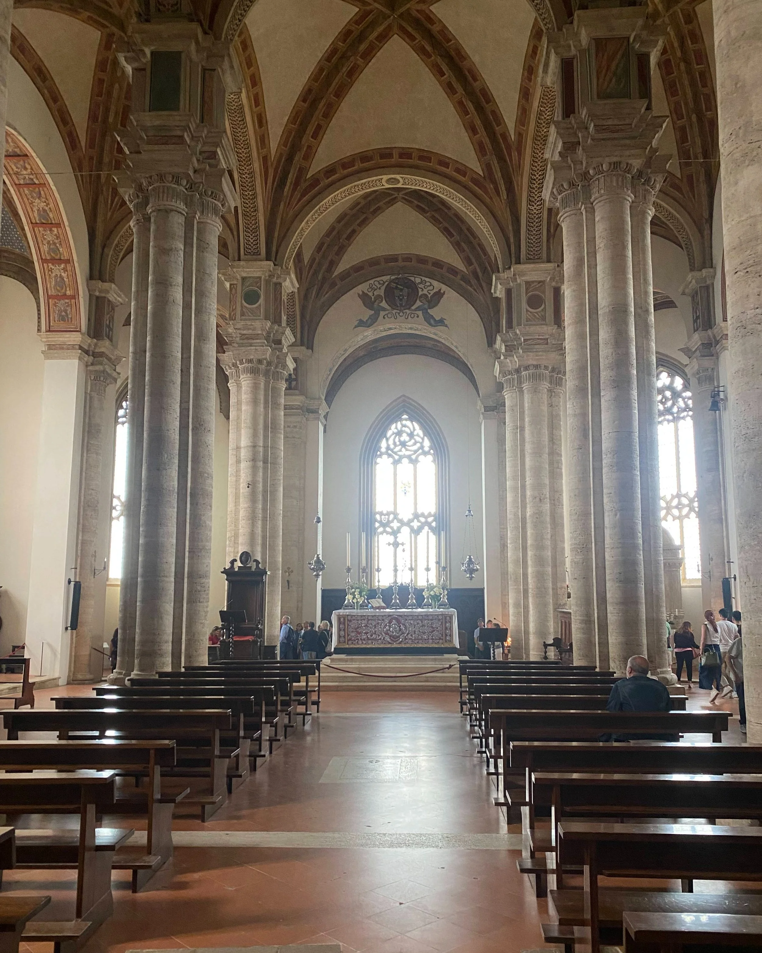 Interior de una iglesia con columnas altas, vitrales grandes y un altar decorado, con algunos feligreses sentados y otros de pie.