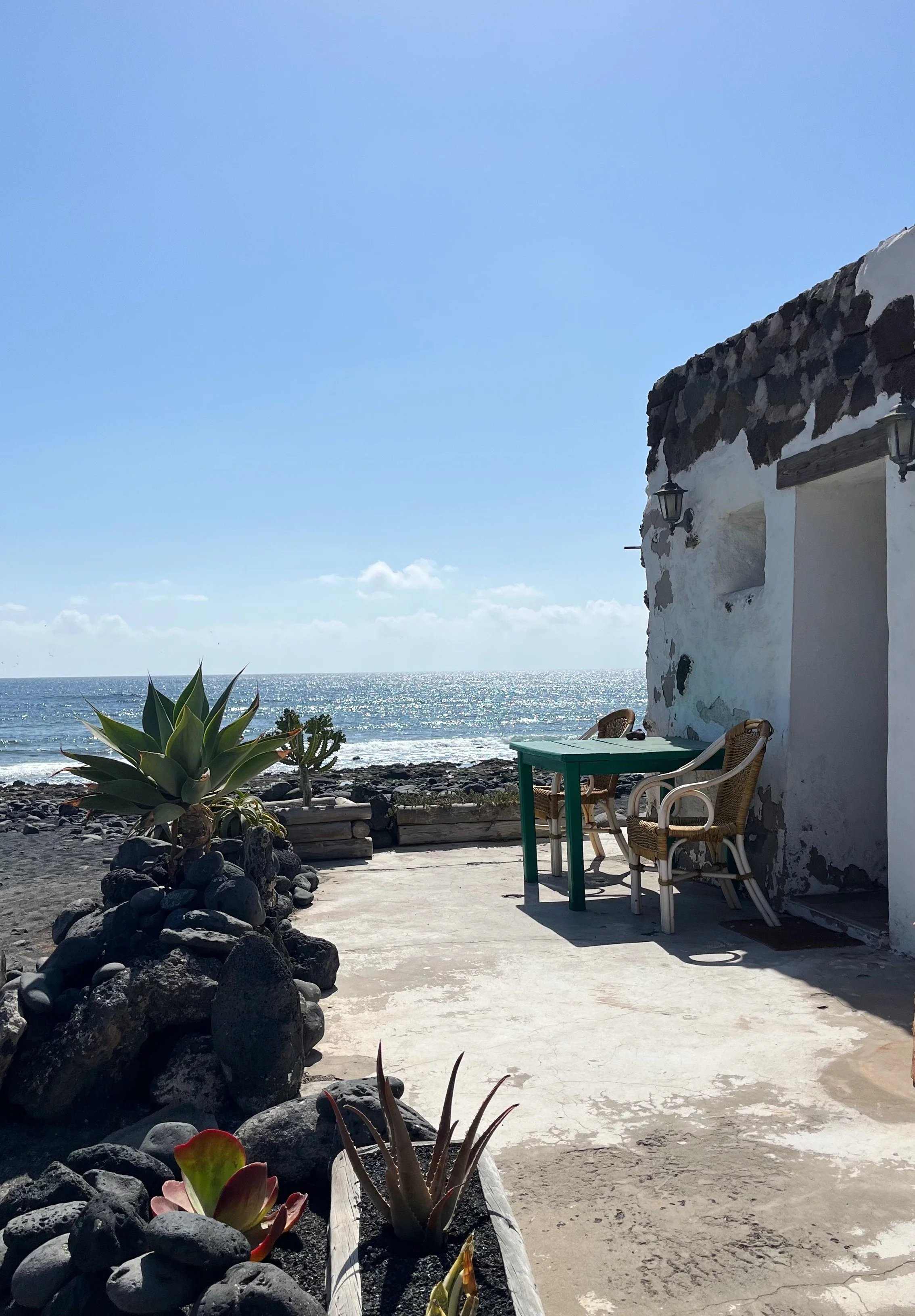 Vista de una terraza junto al mar con plantas suculentas, una mesa verde, dos sillas de mimbre y una casa blanca con piedra en las paredes bajo un cielo azul