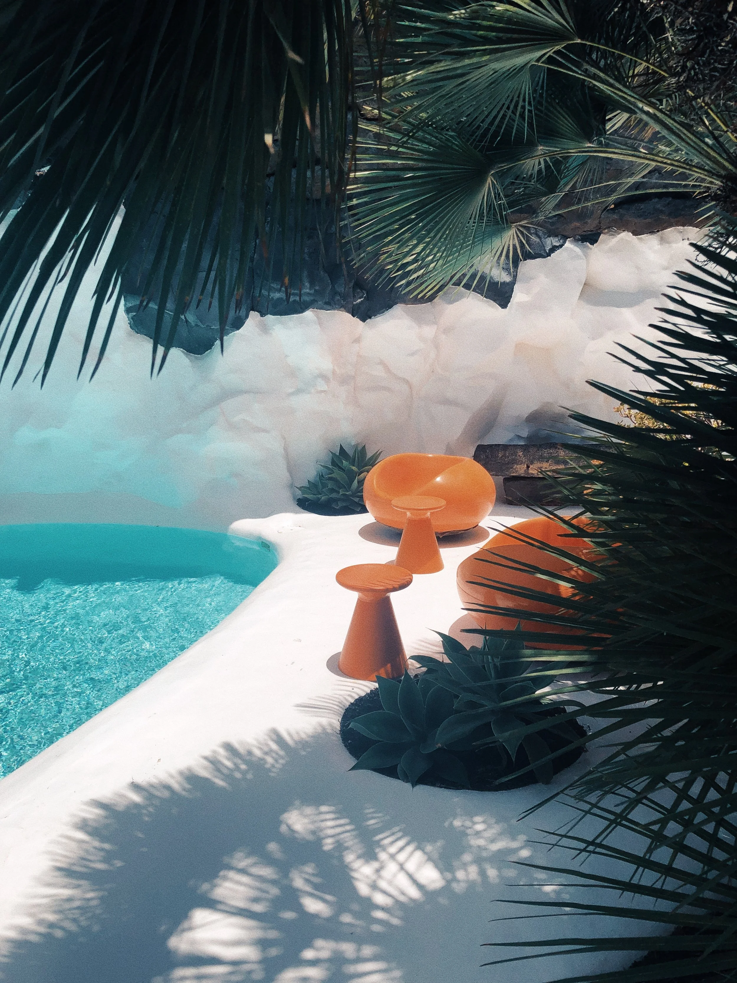 Área de piscina con mobiliario de color naranja, rodeada de plantas tropicales y sombra de hojas.