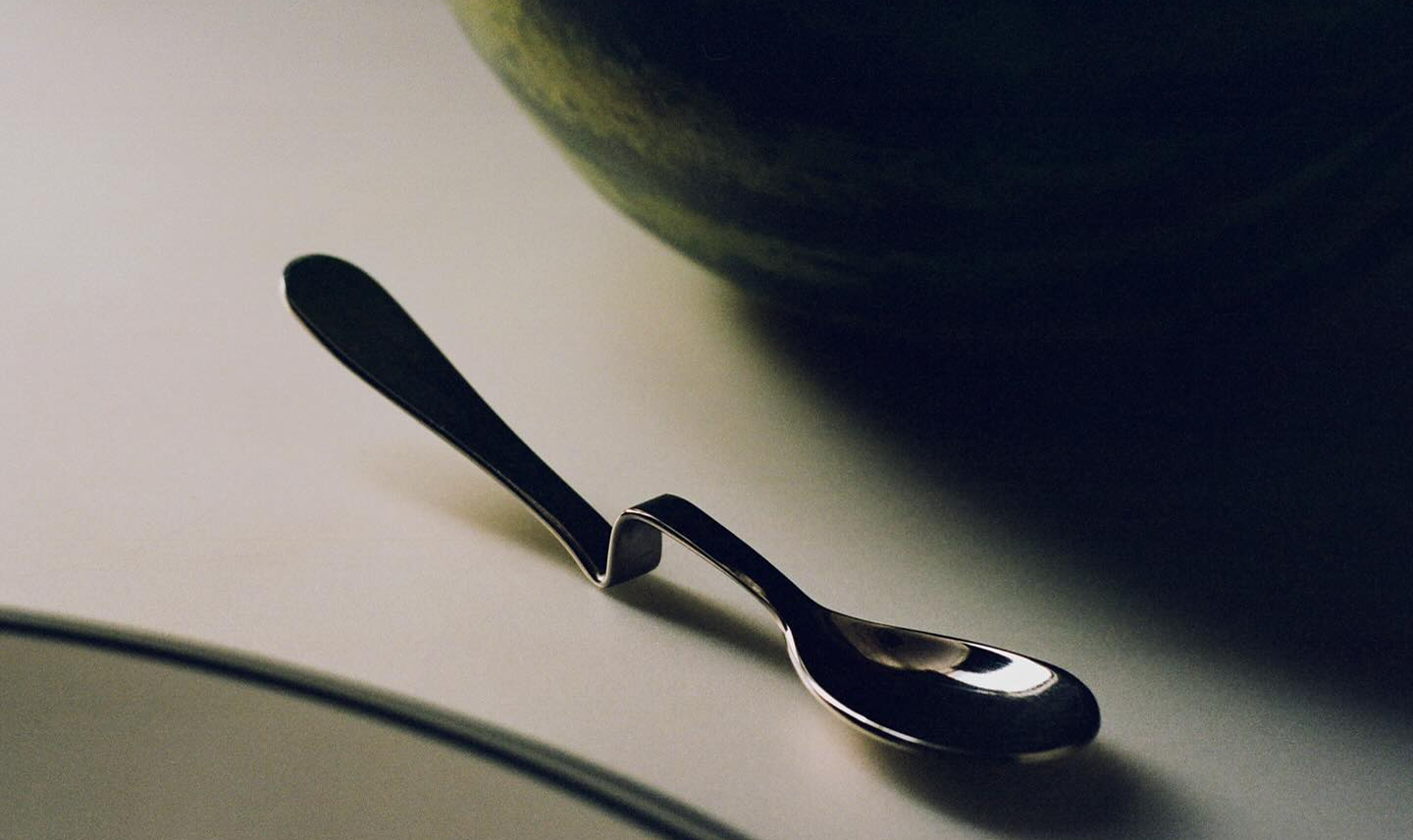 Cuchara de metal en una mesa blanca, parcialmente en sombra, con una sandía grande en el fondo.