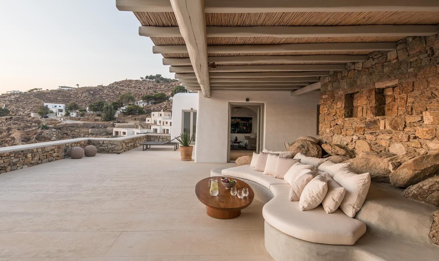 Terraza con sofá de curvas y cojines, mesa de madera con bebidas y frutas, vista hacia casas blancas en colina, decorada con piedra y plantas.