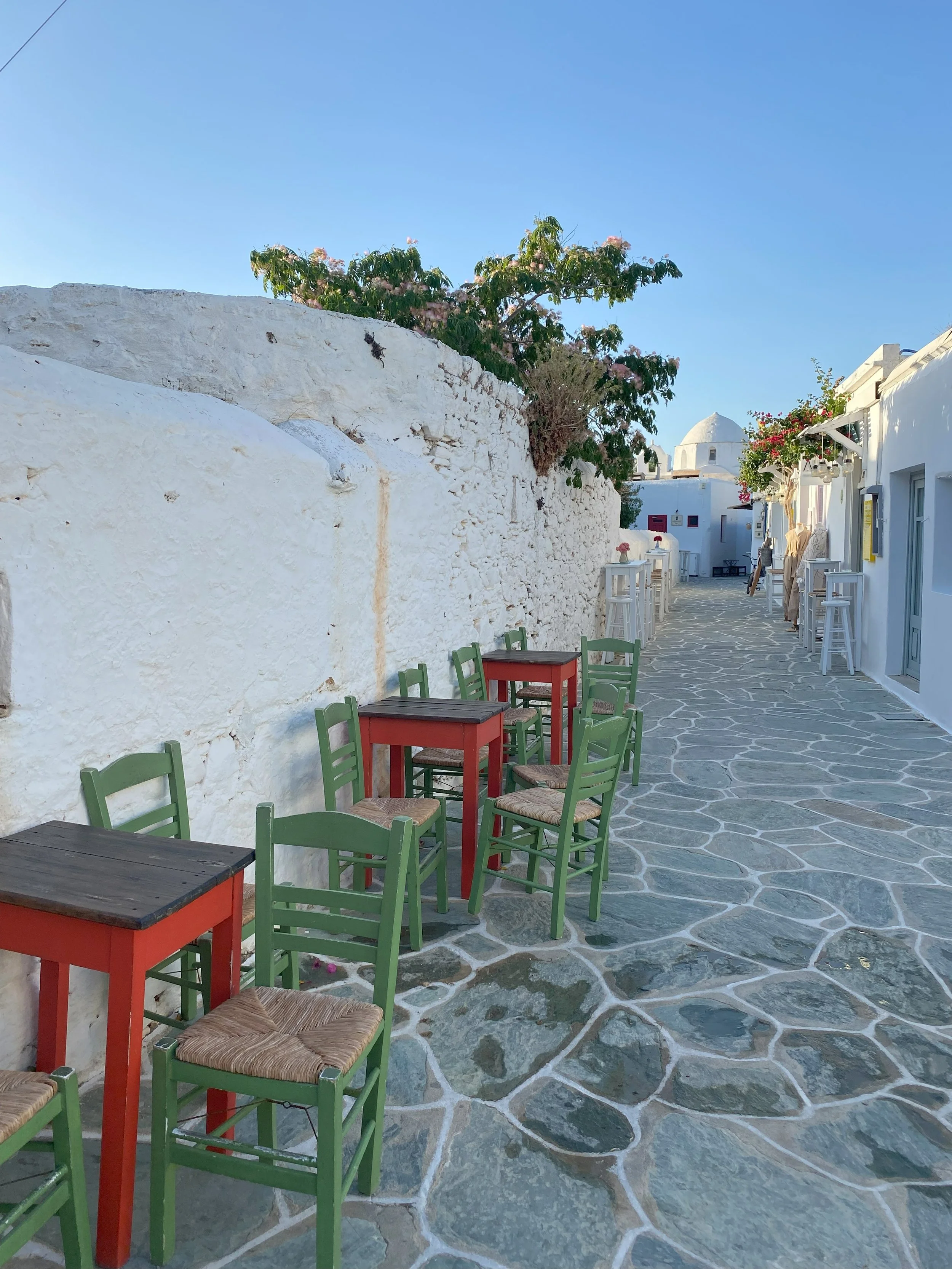 Balcón con mesas y sillas color verde y rojo en un pueblo blanco y soleado, con cielo azul y paredes de piedra