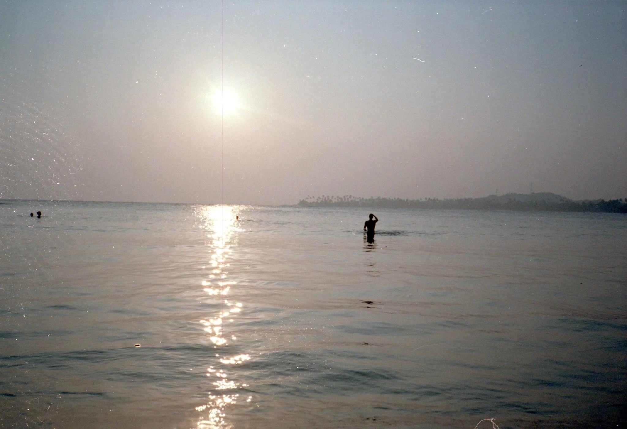 Personas nadando en el mar con el sol brillante en el cielo y un lugar con palmeras en la distancia.