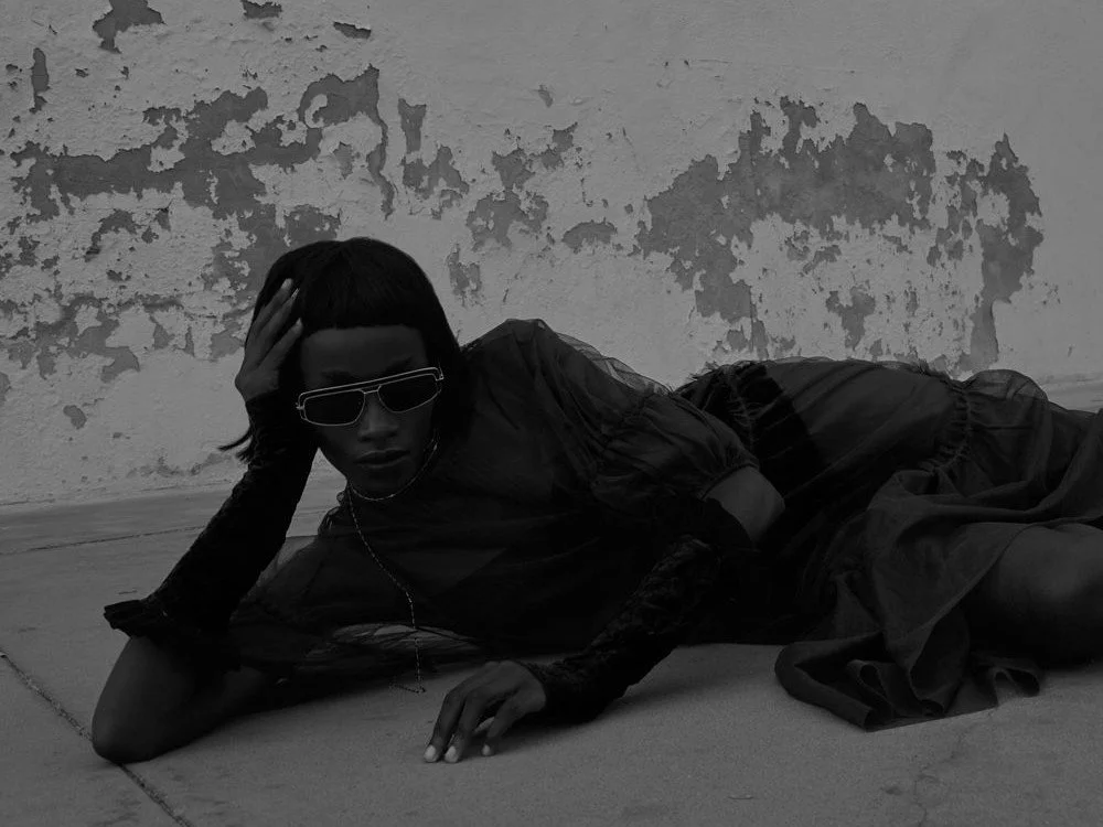 Una persona mujer con gafas oscuras y vestido negro, acostada en el piso, en un fondo de pared con pintura descascarada.