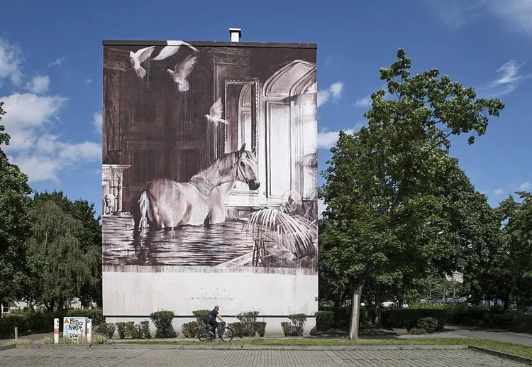 Mural en un edificio que muestra un caballo en un escenario surrealista con palomas en el cielo y elementos arquitectónicos.