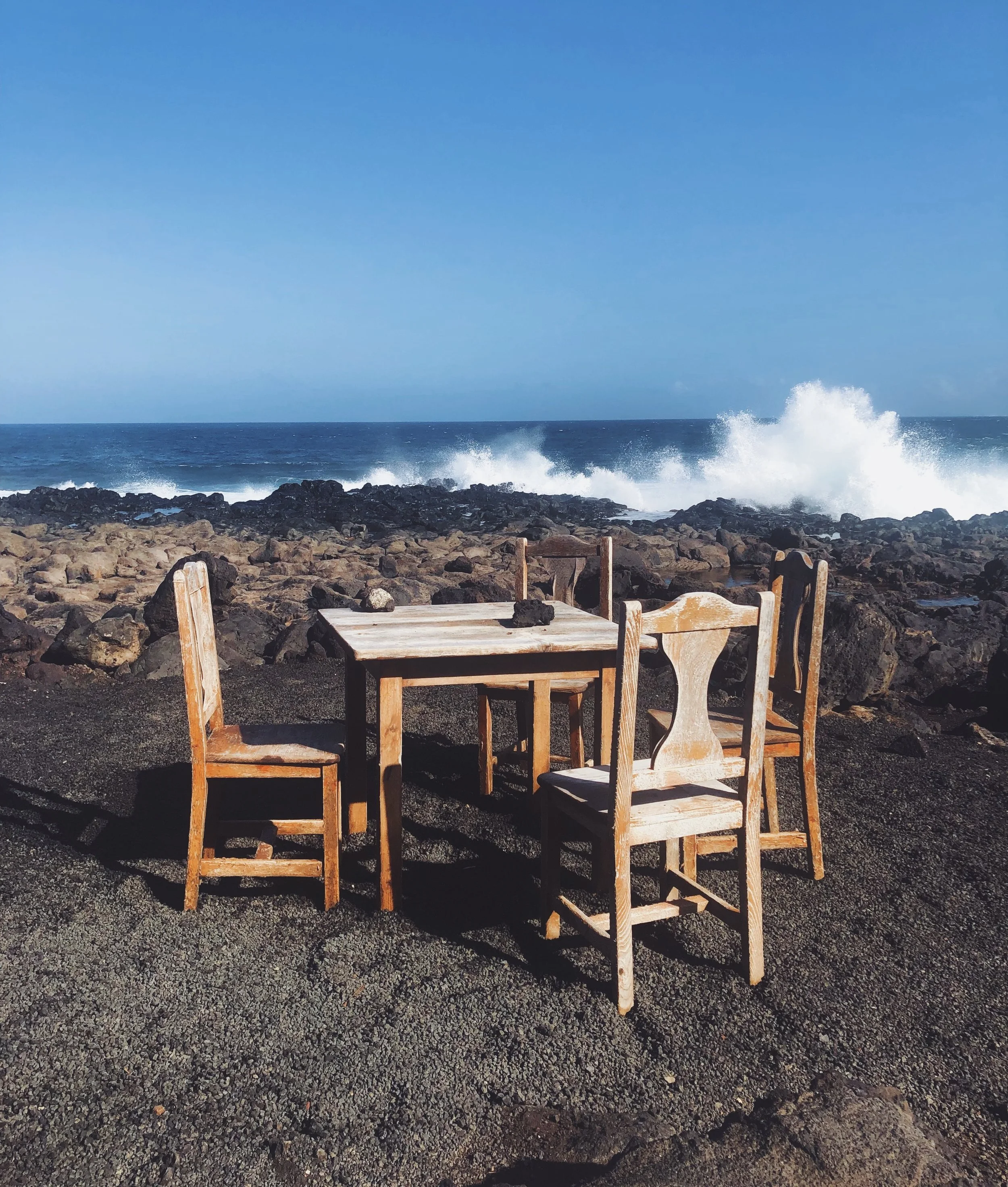 Mesa de madera con cuatro sillas en una zona rocosa frente al océano, con olas rompiendo en la costa y cielo despejado.