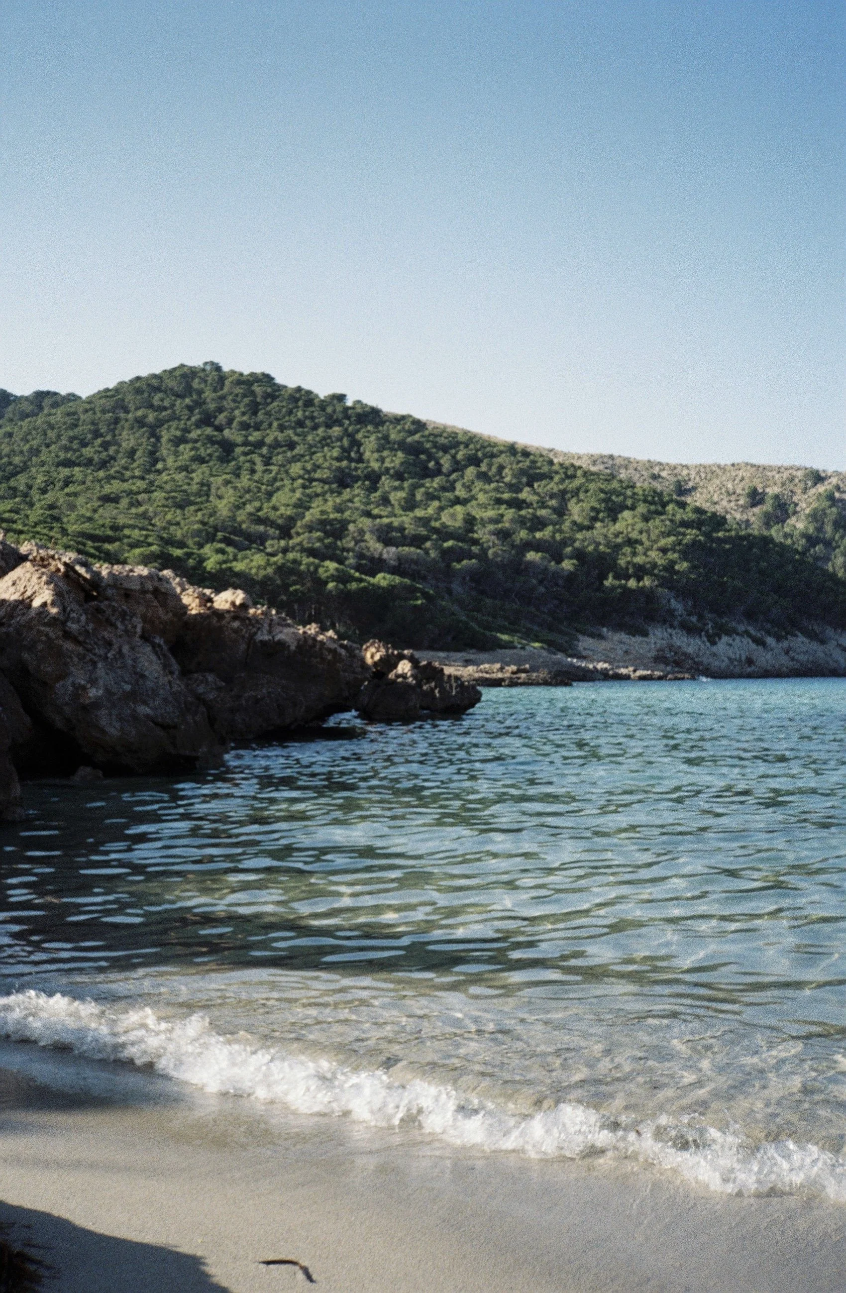 Costa con arena y rocas, agua clara y verde, fondo de colinas con árboles verdes y cielo despejado.