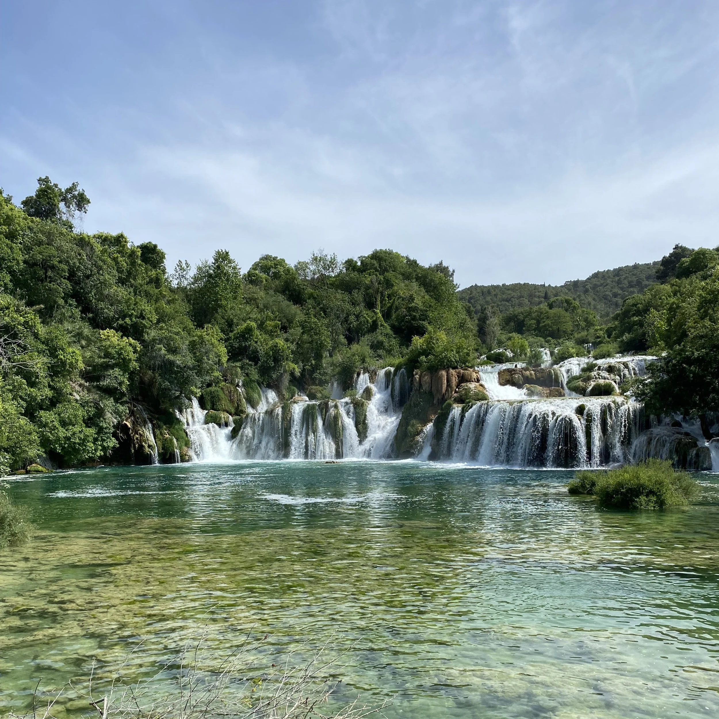 Cascada natural en un río rodeada de vegetación y árboles verdes, con cielo despejado.