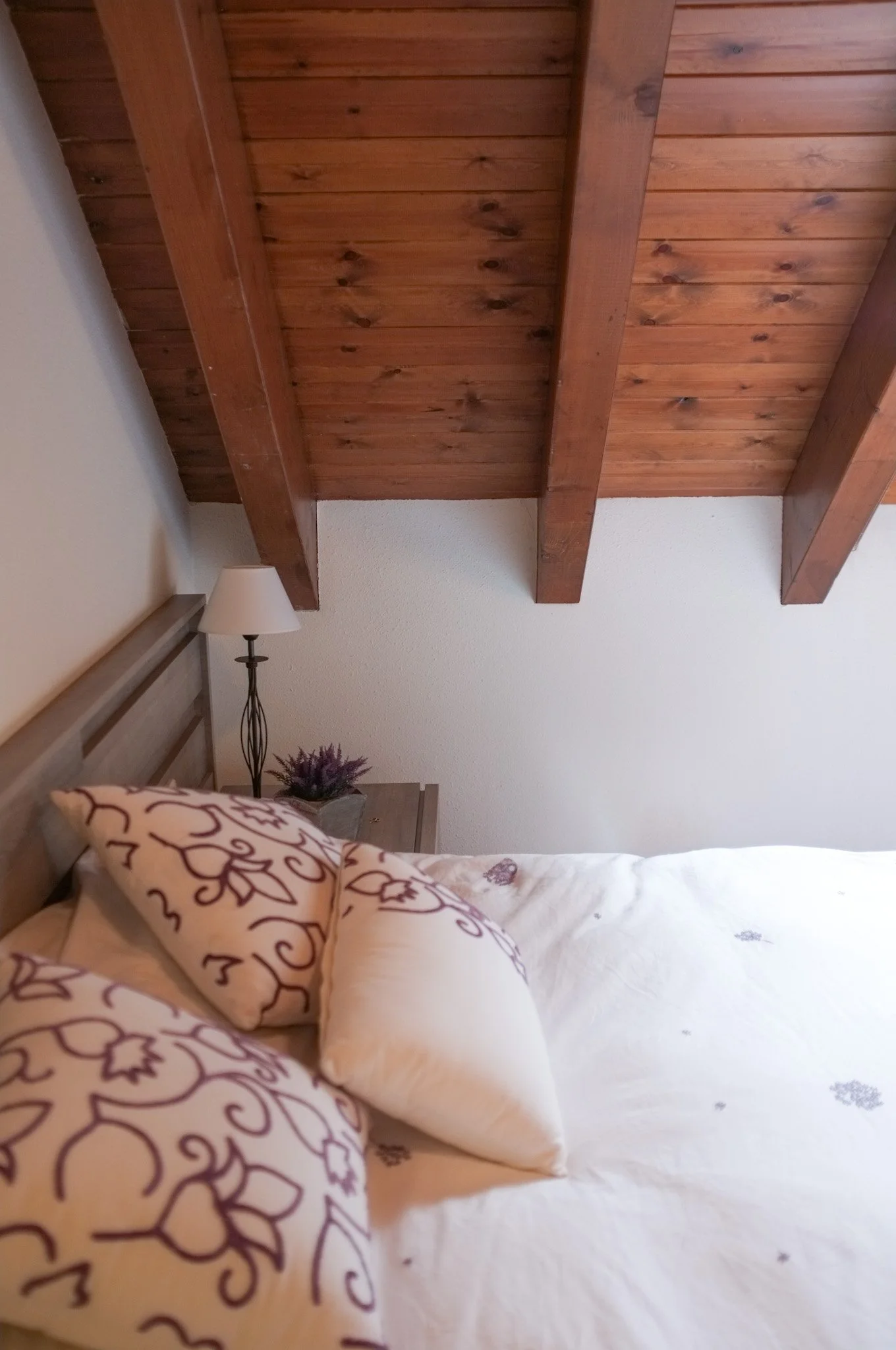 Cama con almohadas decorativas, una lámpara de pie, mesita de noche y techo de madera en una habitación.