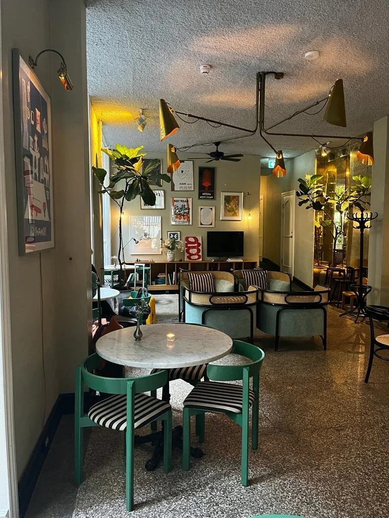 Interior de una sala con varias sillas y sofás, lámparas decorativas, cuadros en la pared, plantas grandes y una televisión