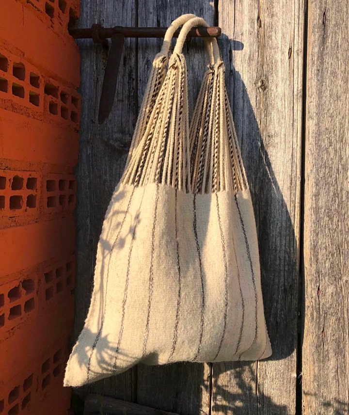 Bolsa de tela tejida de color crema con rayas delgadas negras, colgada en una gancho sobre una pared de madera envejecida, con sombra en la pared y una pared de ladrillos rojos a la izquierda.