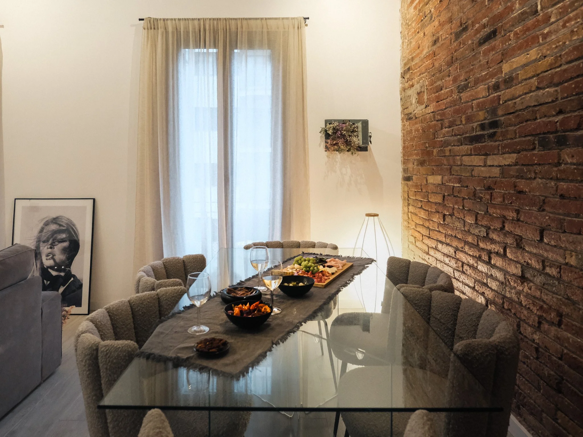 Comedor con mesa de vidrio y sillas beige, decorado con alimentos, vino y una lámpara de pie junto a una pared de ladrillos expuesta, en una sala con cortinas blancas y una obra de arte en el suelo.