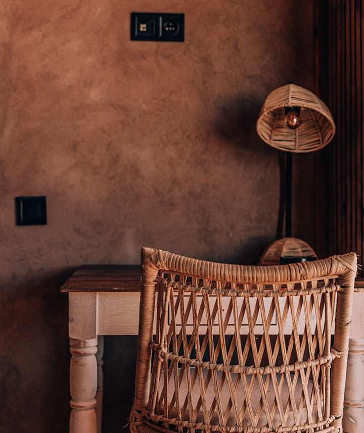 Una mesa de madera con una silla de respaldo de mimbre en una habitación con pared de color marrón y una lámpara de piso de mimbre