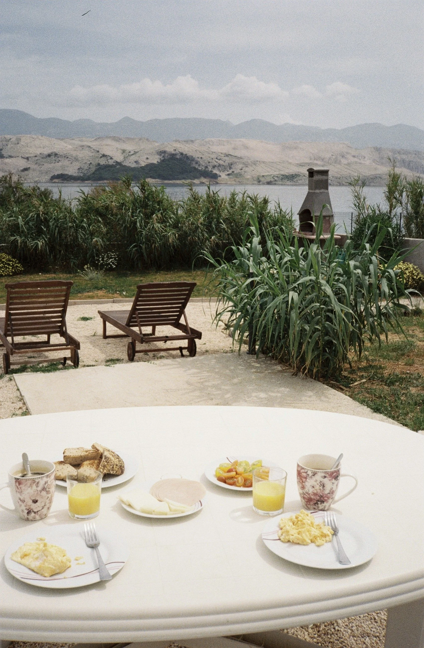 Mesa con desayuno, incluyendo huevos, pan, jugo, queso, y taza de café o té, en un patio con vista a un lago y montañas.