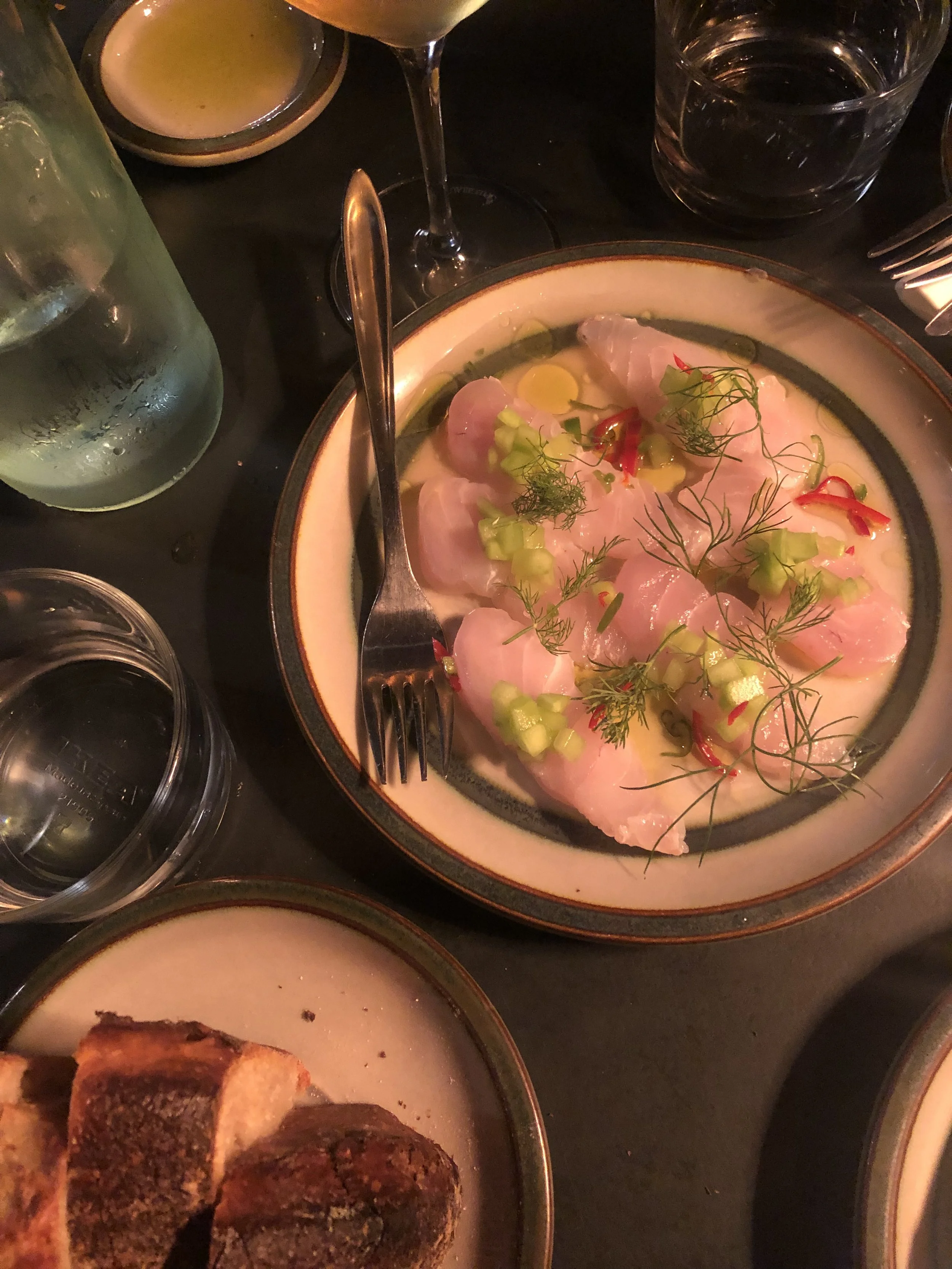 Plato de ceviche de pescado decorado con cilantro, rodajas de chile y cebollín, acompañado de pan tostado y varias bebidas como agua, vino y agua con hielo.