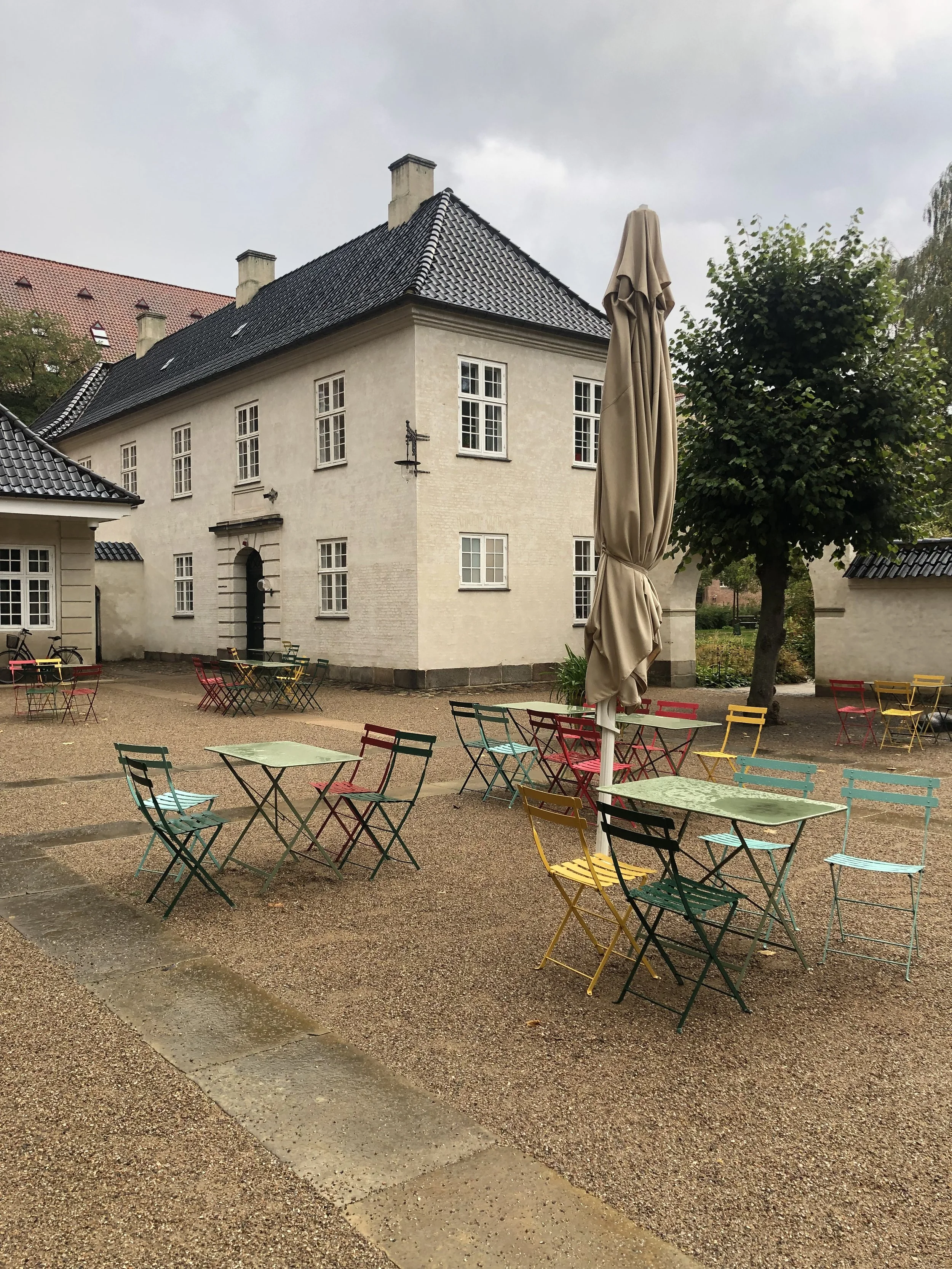 Terraza con mesas y sillas de colores en un patio frente a un edificio blanco con ventanas y techo de tejas negras, en un día nublado.