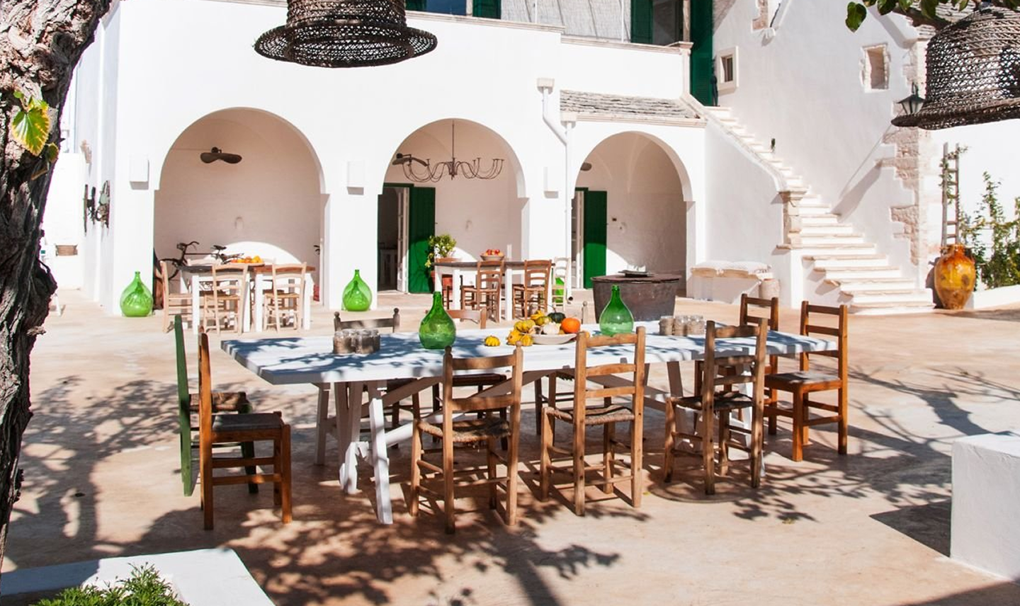 Vista de un patio al aire libre con mesas y sillas de madera, decoradas con jarrones verdes y un ambiente de estilo mediterráneo con paredes blancas y escaleras.