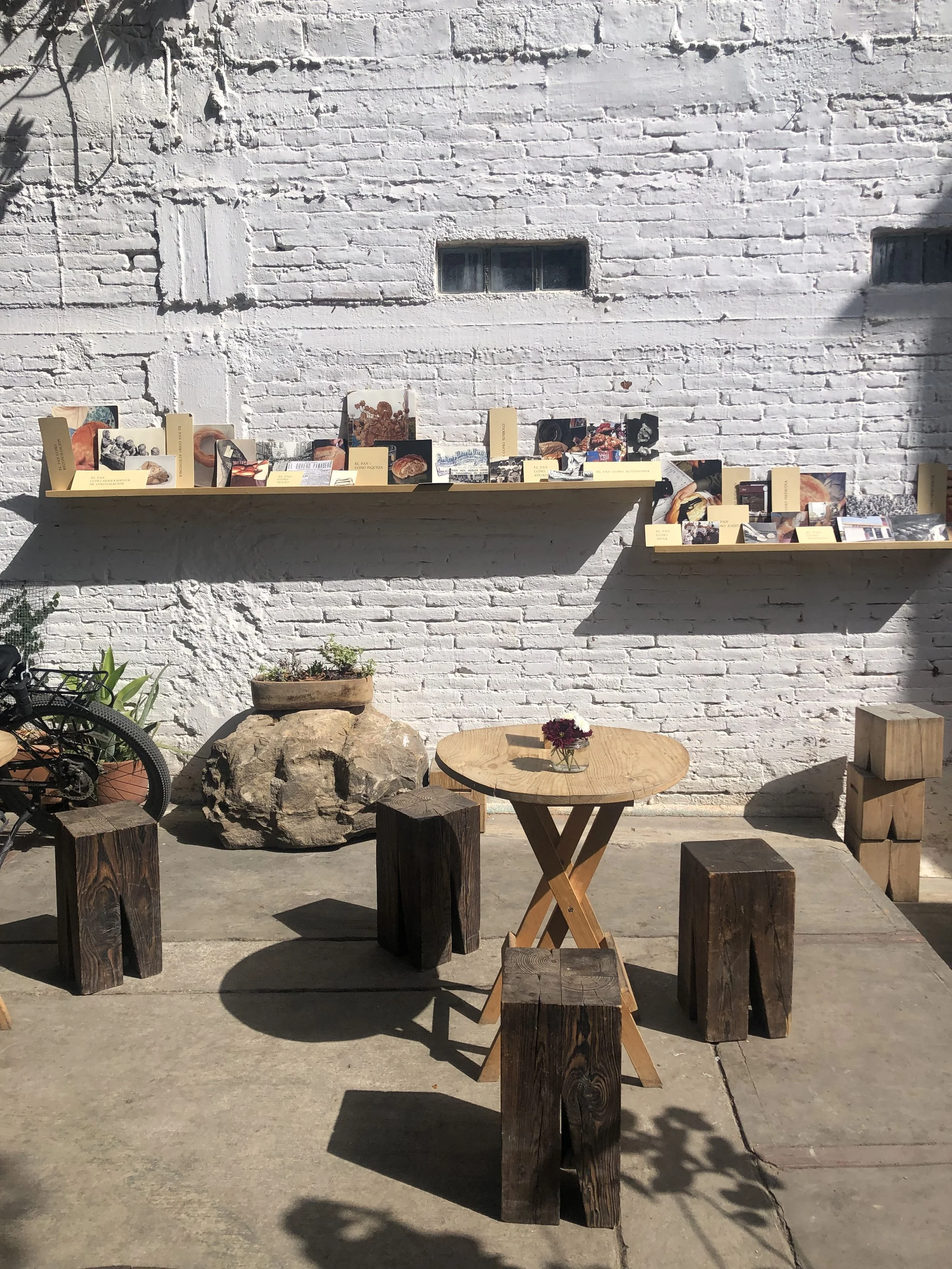 Mesas de madera y bancos en un espacio al aire libre con pared de ladrillos blancos y estantes con fotografías y notas.