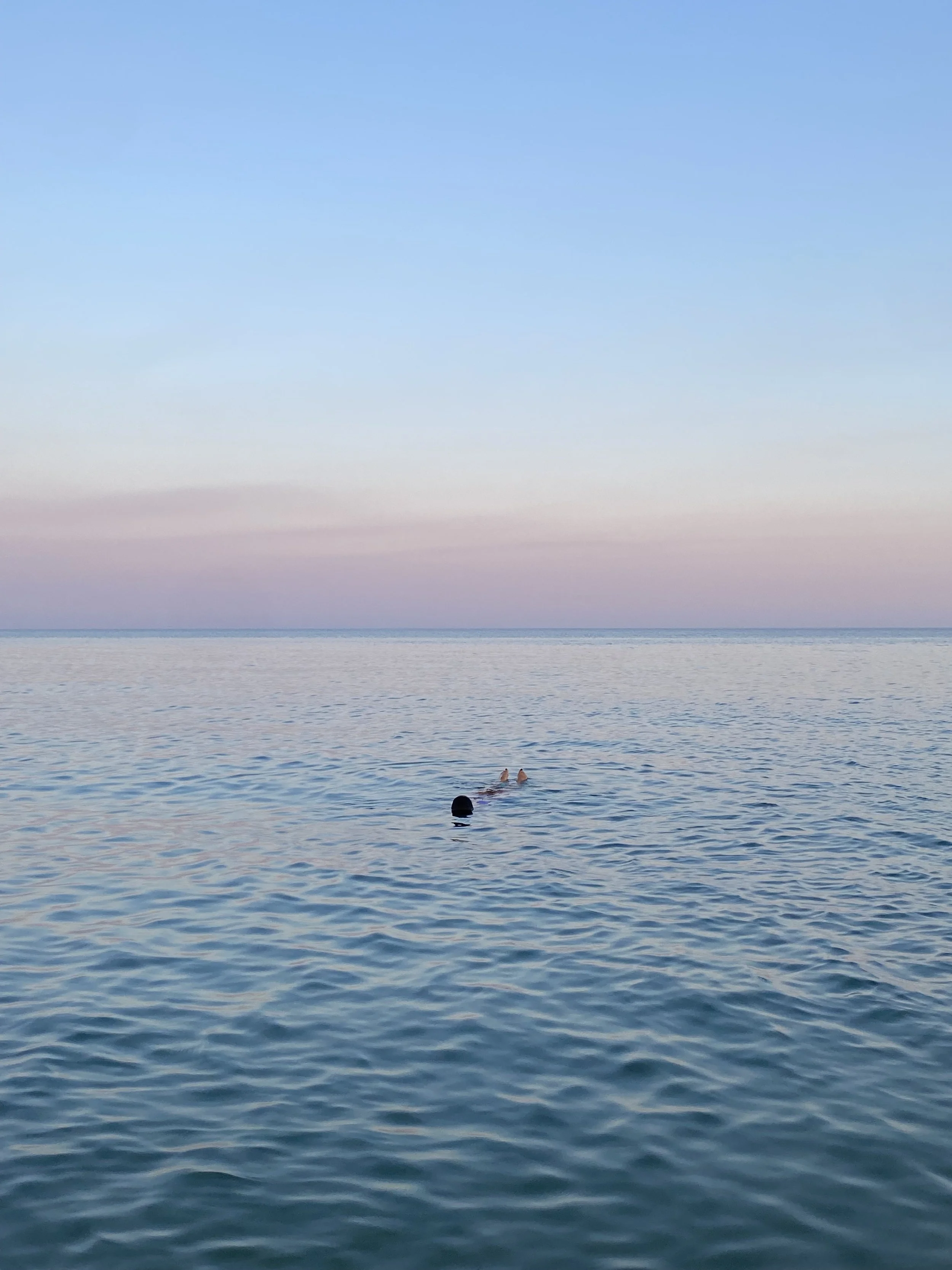 Mar en calma con un cielo a tonos pastel y dos personas nadando en el mar.