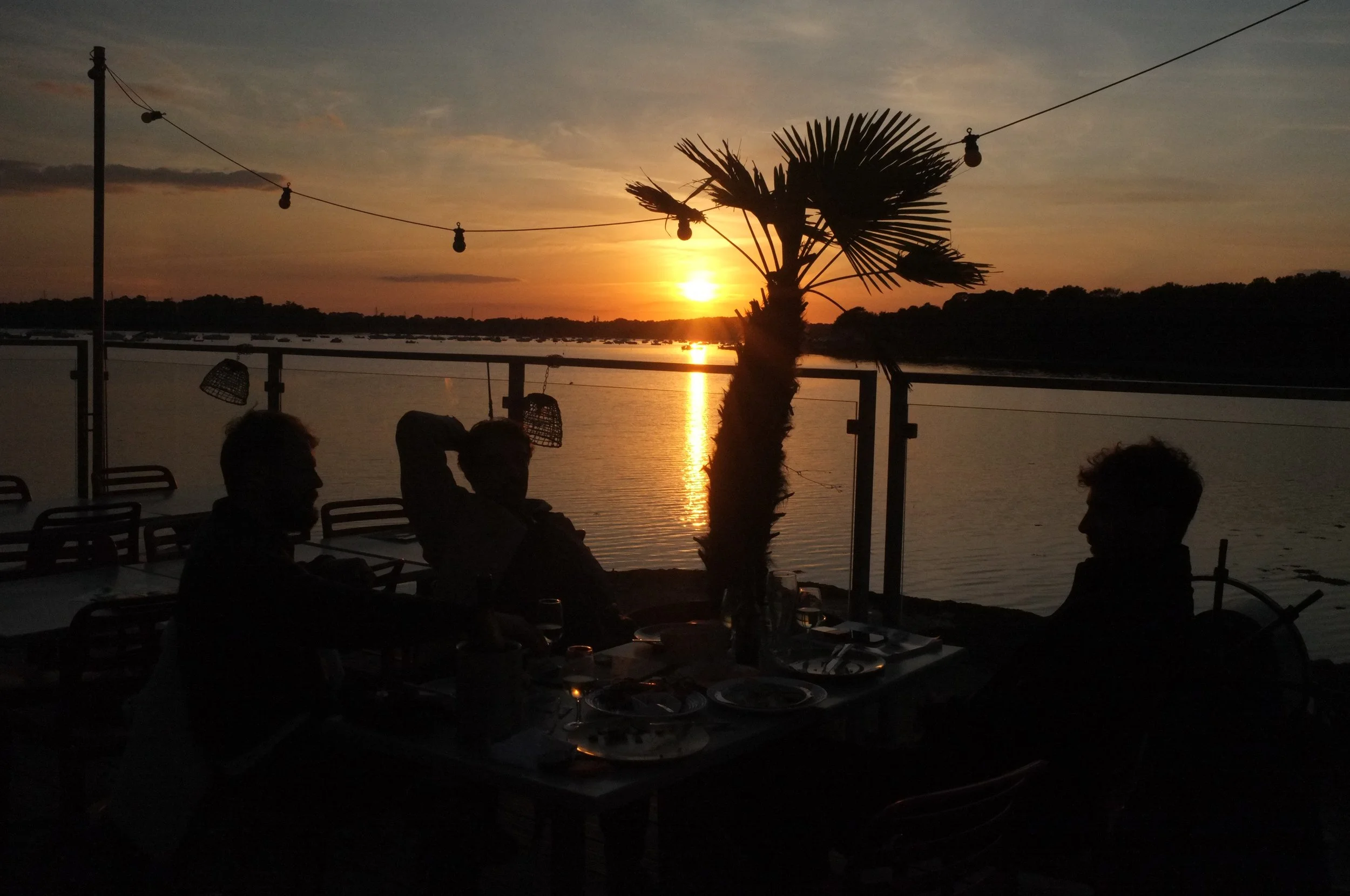 Personas cenando en un restaurante al aire libre durante la puesta del sol, con vista a un río y una palmera en el fondo, decoraciones de luces colgantes.