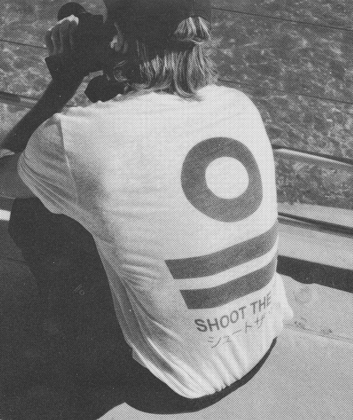 Persona pescando desde un barco en el agua, vista de espaldas, usando una camiseta con un diseño y texto que dice 'Shoot the'.