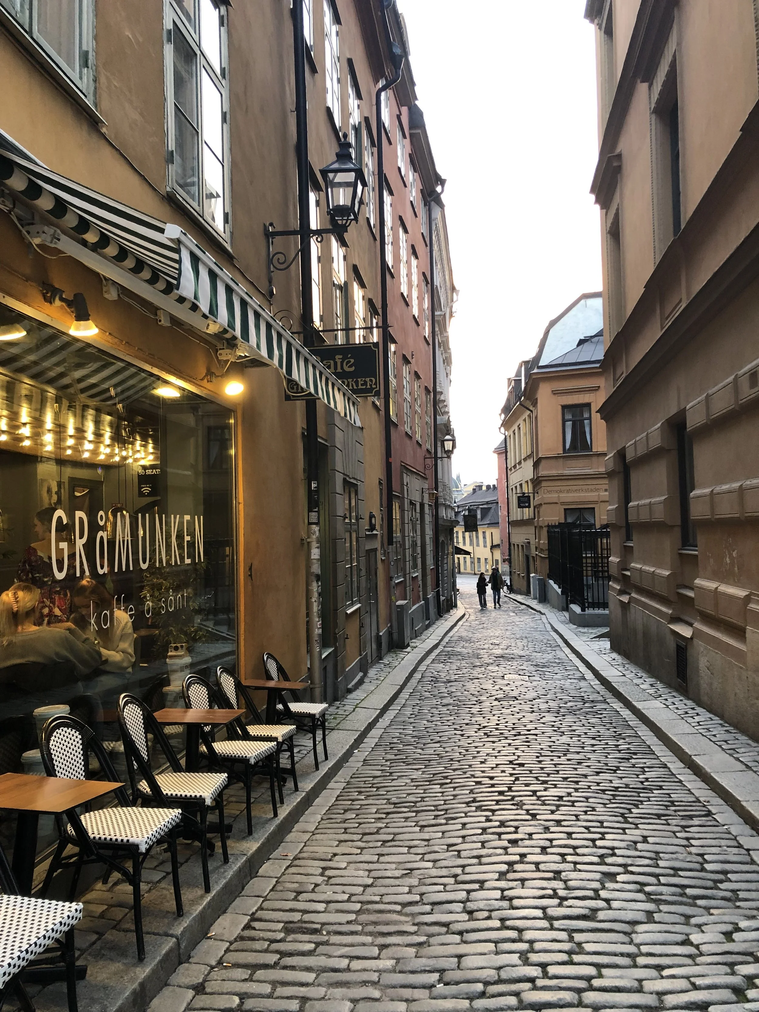 Calles empedradas en una ciudad europea con edificios coloridos, un café con sillas en la acera y personas caminando en la distancia, en horario de atardecer.