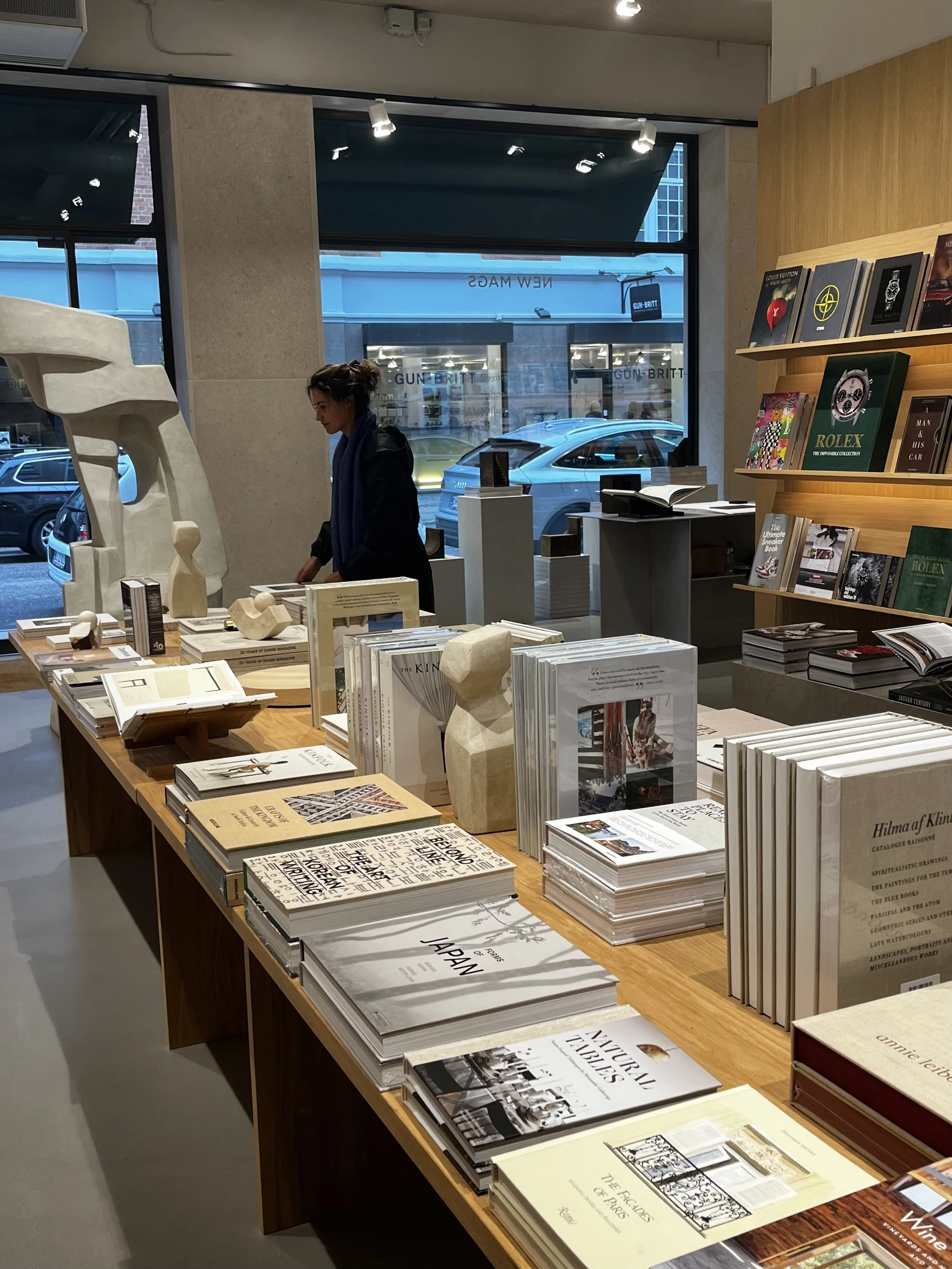 Interior de una librería con mesas llenas de libros y una mujer examinando uno en la mesa