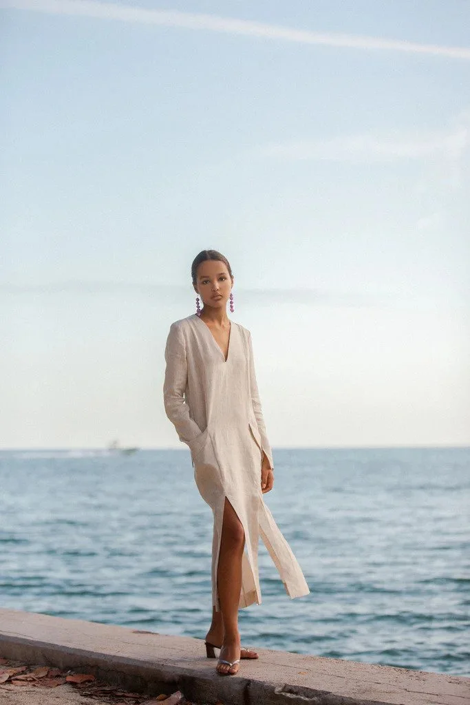 Una mujer de pie en un muelle junto al mar, usando un vestido largo beige y pendientes grandes, con el mar y el cielo de fondo.